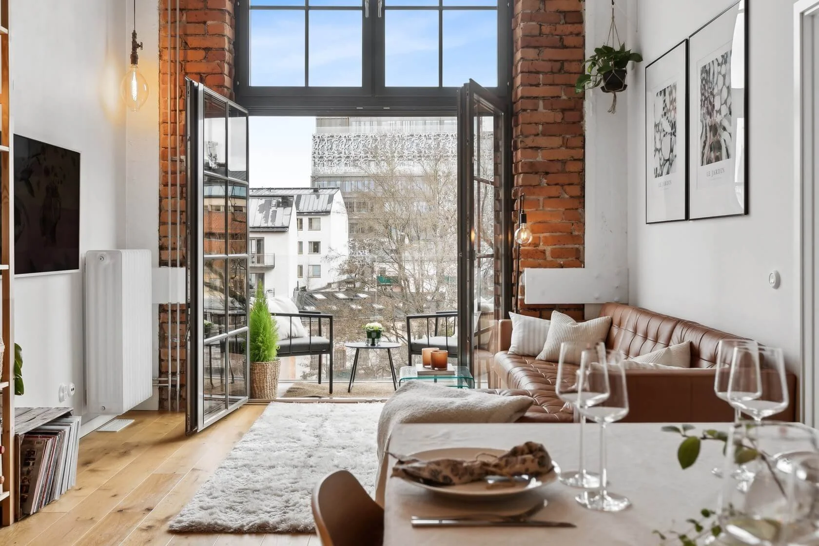 Living room with exposed brick wall, white walls, and large windows opening to a balcony with city views. Features a brown leather sofa with pillows, a white fluffy rug, a small coffee table with plants and candles, and a dining table set with wine g