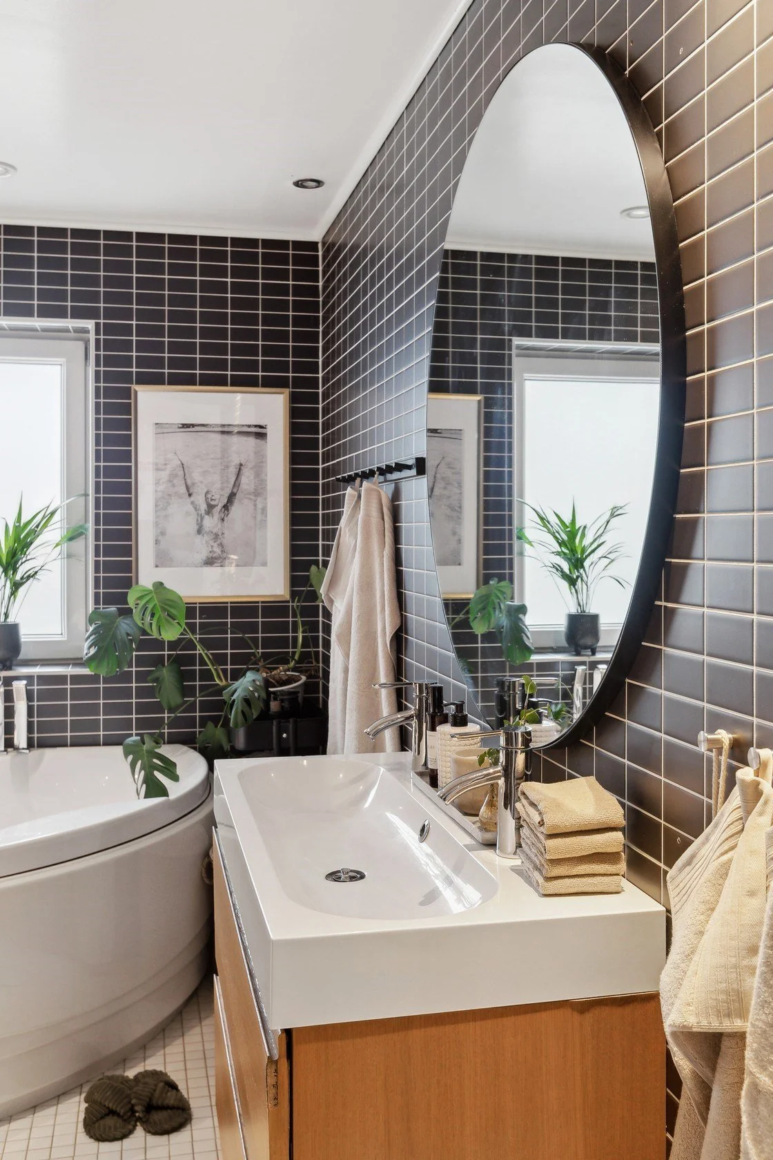 A modern bathroom with a black tile wall, a large mirror, a white sink with a wooden cabinet, beige towels, plants, and framed black and white photos.