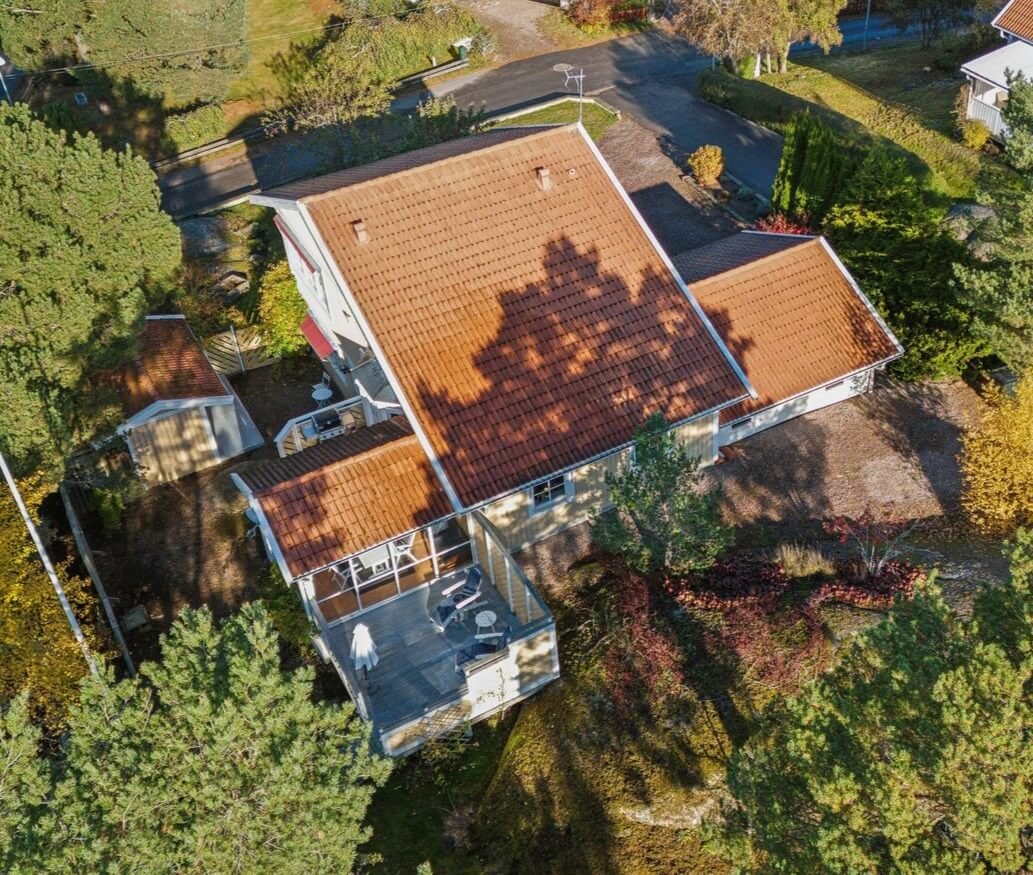 An aerial view of a house with a red tiled roof surrounded by trees and shrubbery. There is a deck with outdoor furniture and umbrellas attached to the house. The house is located near a curved street in a suburban neighborhood.