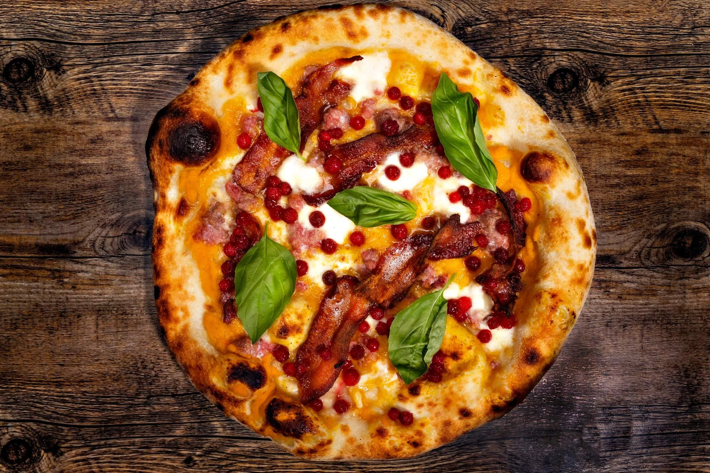 A pizza with bacon, red berries, cheese, and fresh basil leaves on a wooden surface.