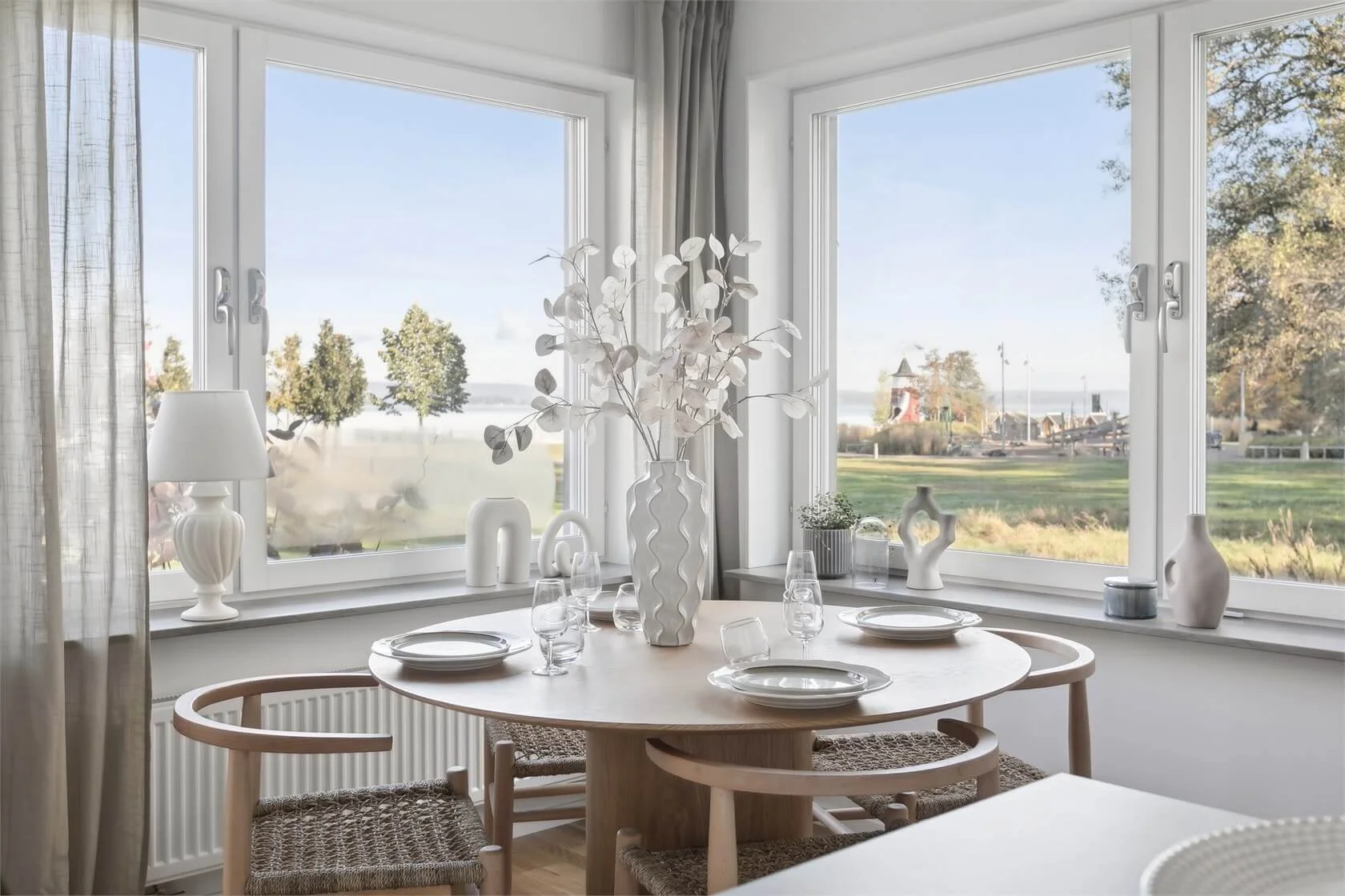 Dining area with round wooden table set with white plates and glasses, surrounded by four woven chairs, in front of large windows showing outdoors with trees and a clear sky.