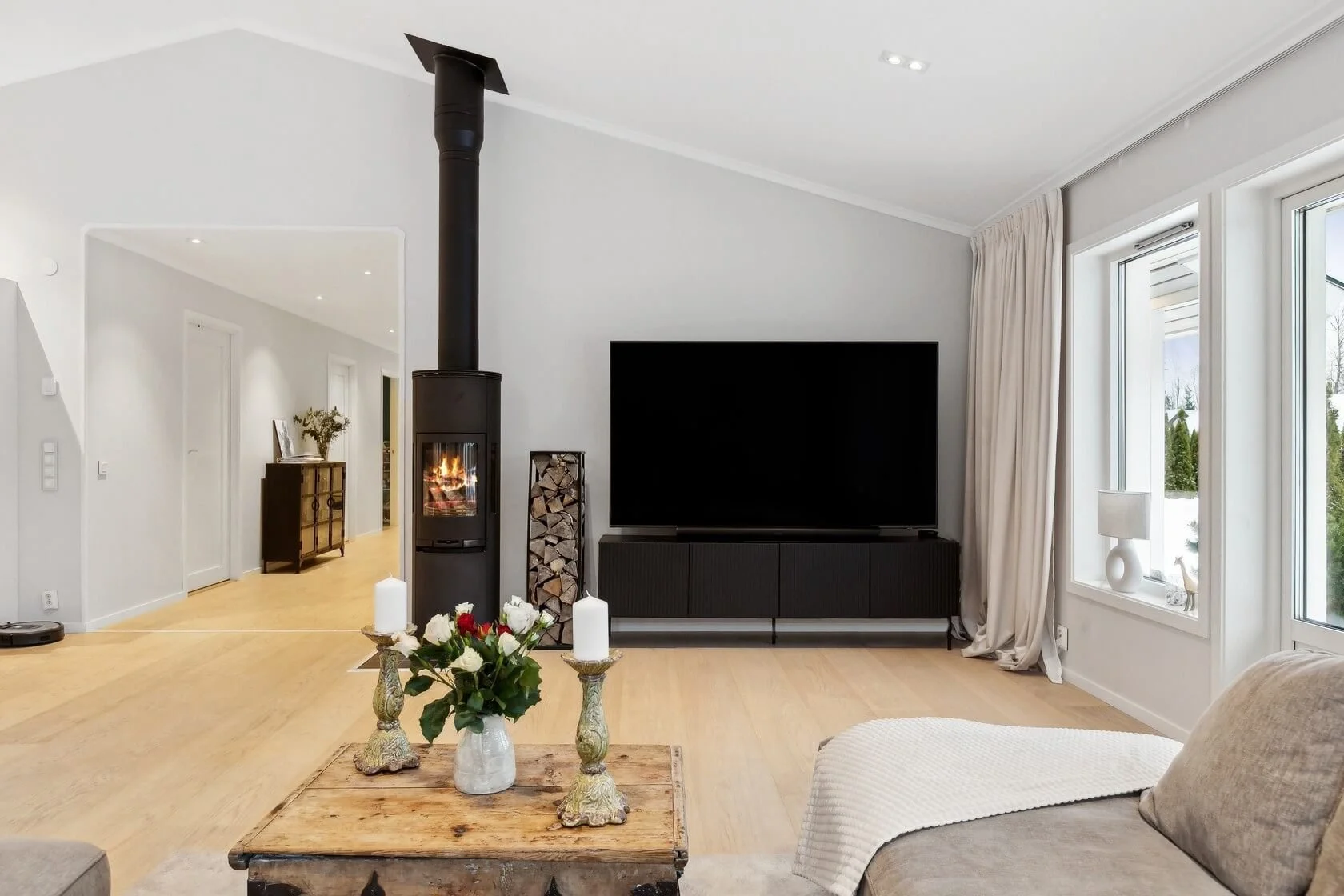 Living room with a black wood stove, flat-screen TV, window with curtains, wooden coffee table, candles, and a beige sofa.