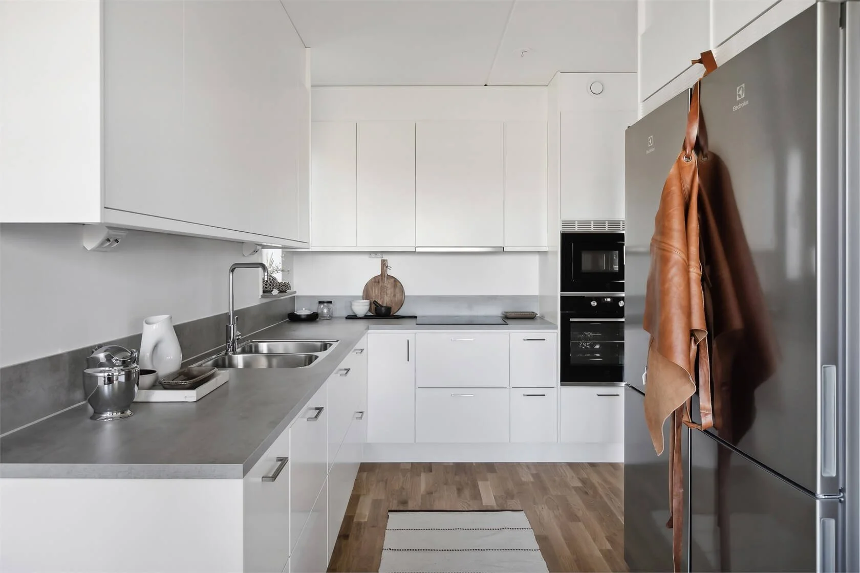 Modern kitchen featuring white cabinets, a gray countertop, a stainless steel sink, and a large refrigerator with brown leather and fabric hanging on it. There is a built-in oven and microwave, wooden flooring, and a small striped rug on the floor.