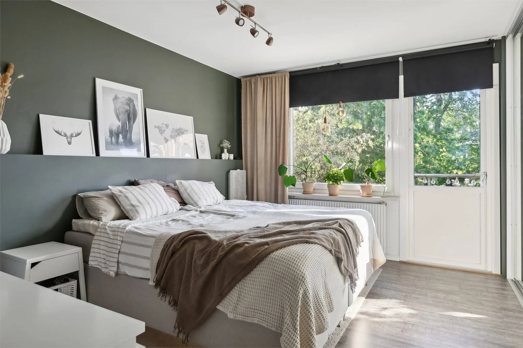 A contemporary bedroom with a green accent wall, white bed with striped pillows, beige throw blanket, framed nature prints above the bed, a window with potted plants, and a sliding glass door leading outside.