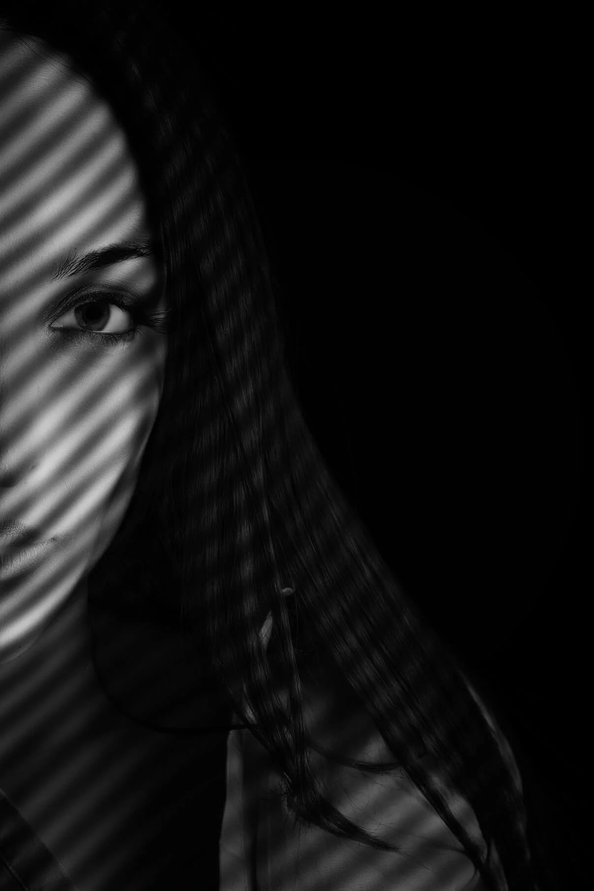 Close-up black and white photo of a woman with one eye visible, partially covered by diagonal blinds, with dark hair and a focused gaze.
