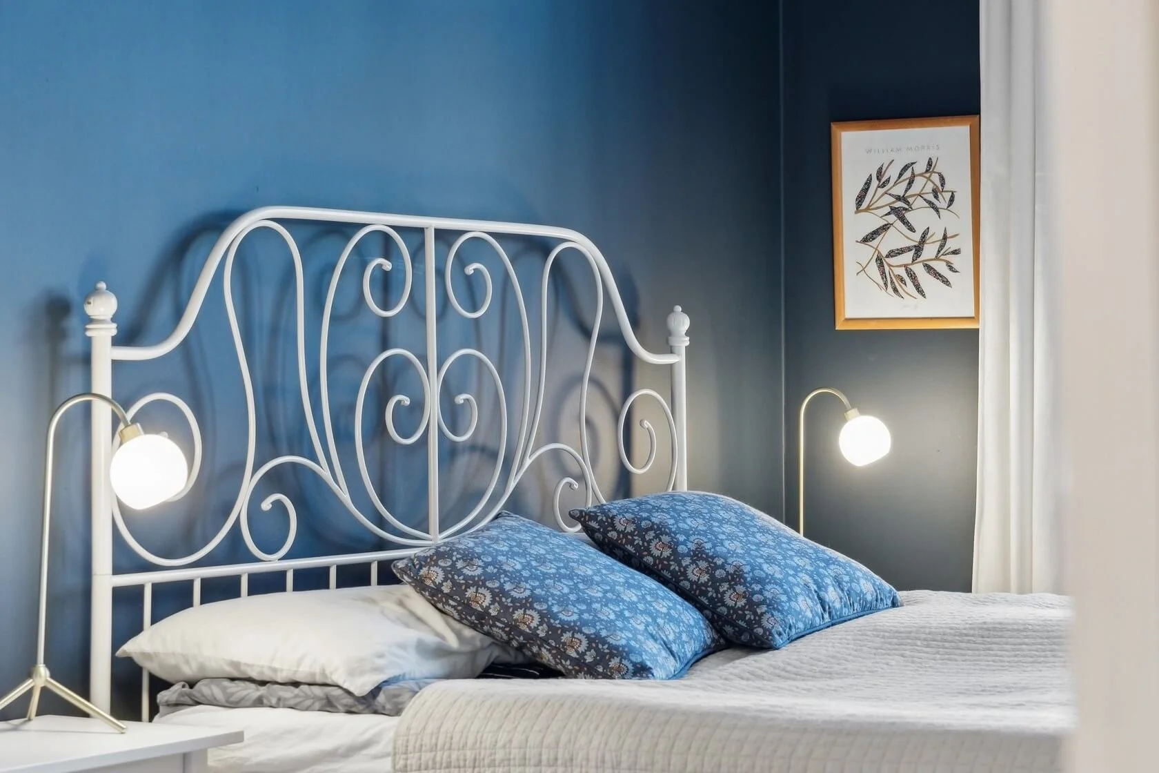 A cozy bedroom with a white metal bed frame, blue floral pillows, white sheet, blue painted wall, framed botanical artwork, white nightstand, and two small wall-mounted lamps with white shades.