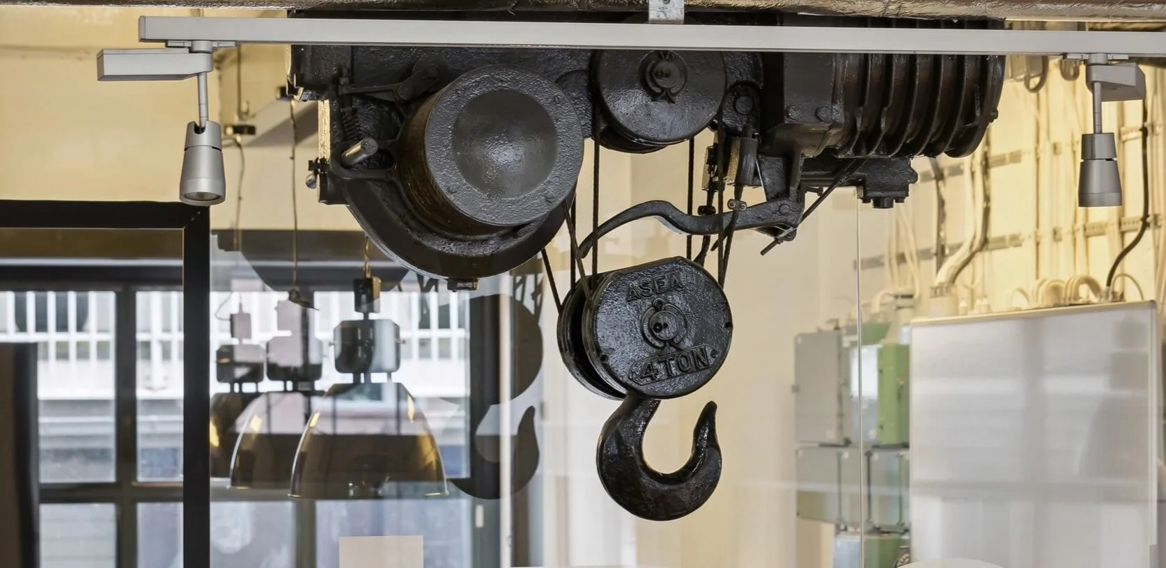 Industrial overhead crane hook and pulley system with black weight plates marked 4 TONS, installed in an indoor setting with large windows and office equipment in the background.