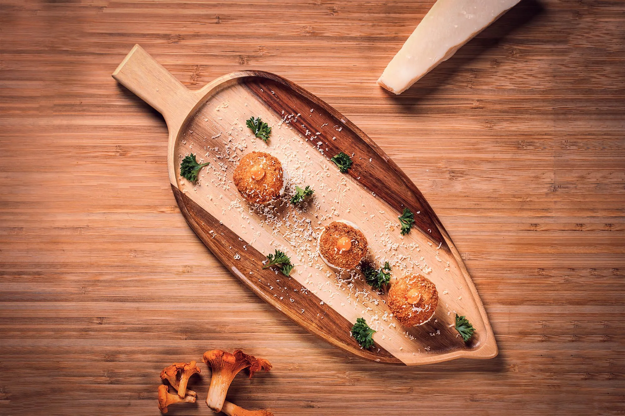 Three croquettes garnished with parsley and grated cheese on a wooden serving tray, with a wedge of cheese and some mushrooms nearby.