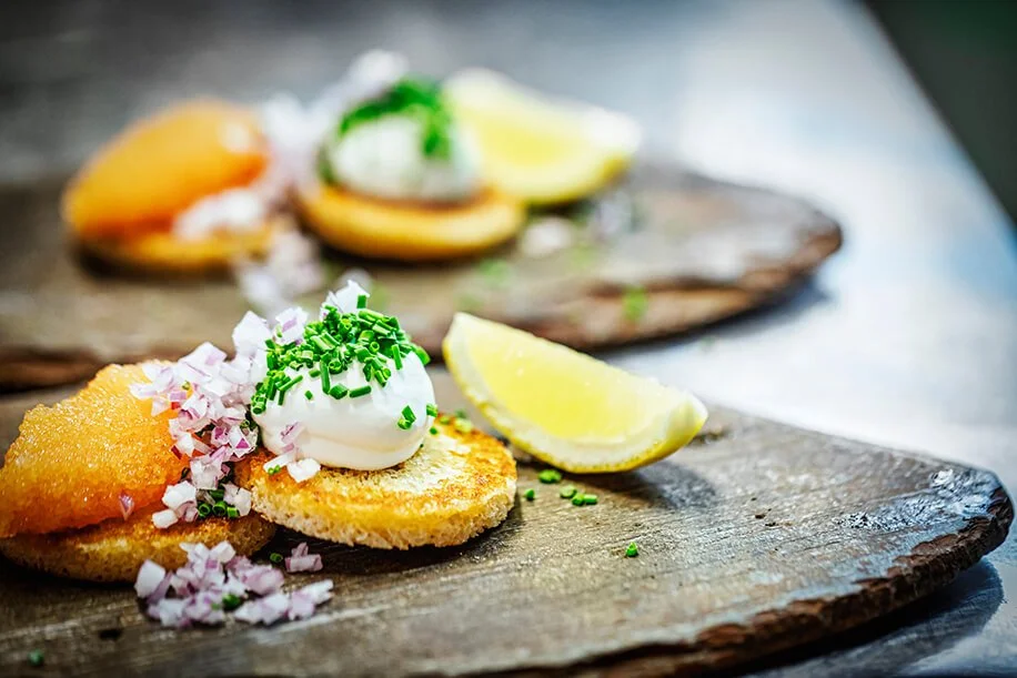 Close-up of two pieces of fried fish, tartar sauce with green sprinkles, lemon wedge, and chopped onion on a wooden serving board
