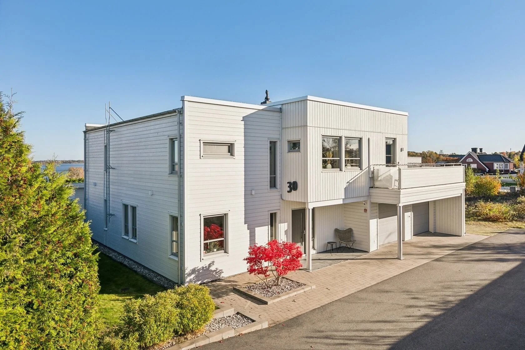 White modern two-story house with a small red tree in front and a driveway on the right side, under a clear blue sky.