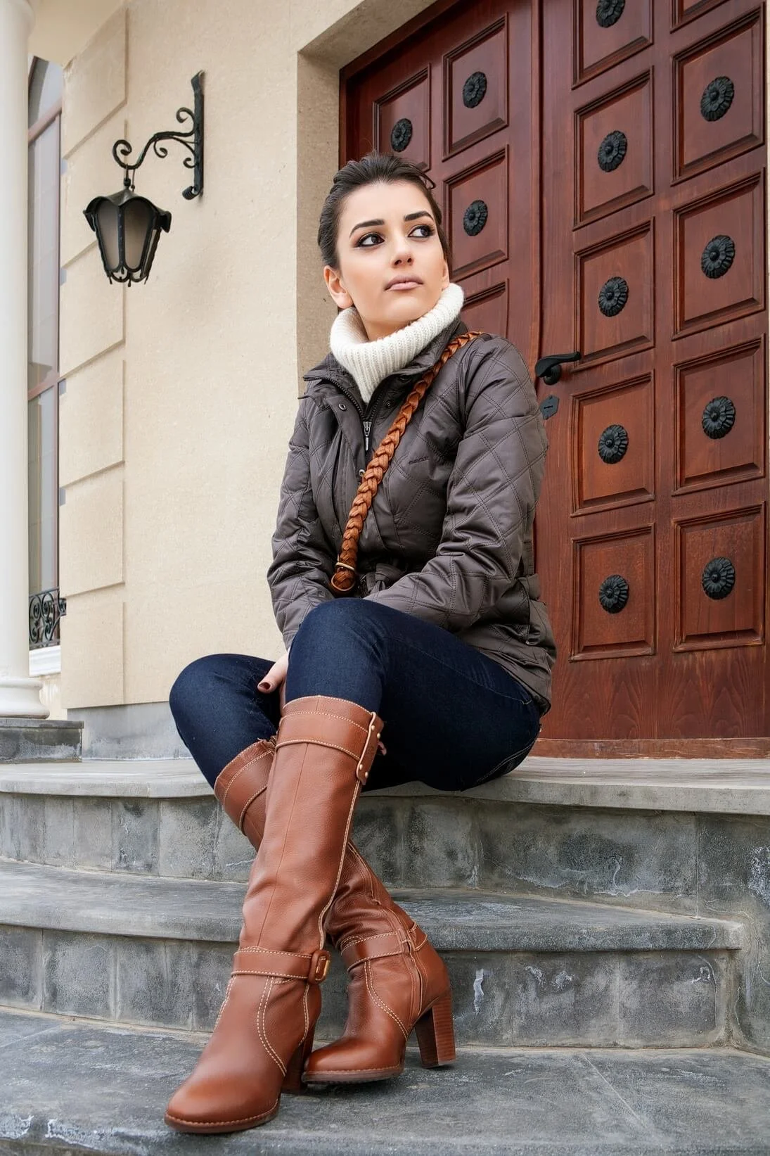 A woman sitting on stone steps outside a house with a wooden door, wearing brown high-heeled boots, dark jeans, a quilted jacket, and a white turtleneck sweater.