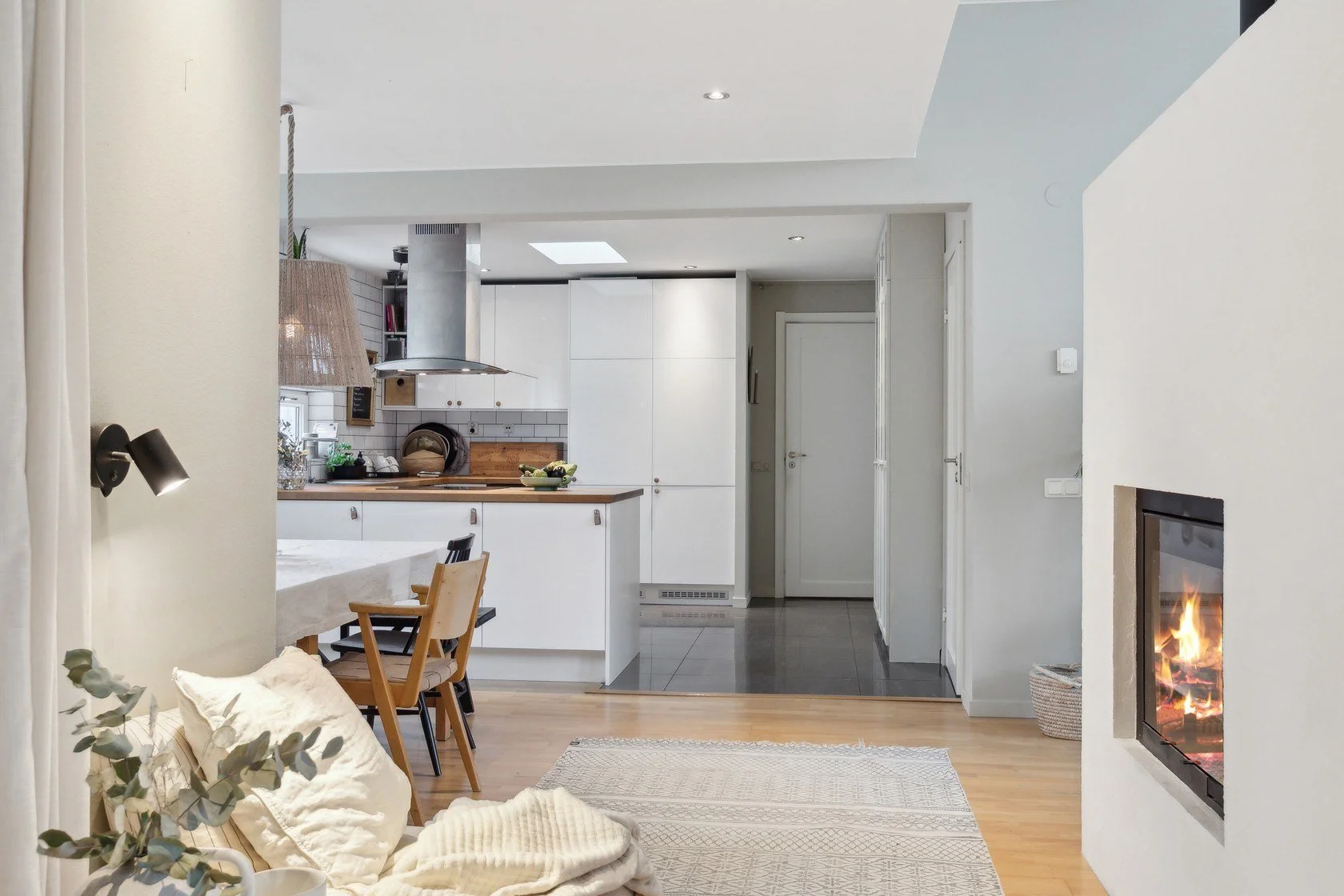 Interior of a modern living space with a fireplace on the right, a dining table with chairs, and a view into a kitchen with white cabinets and a wooden countertop.