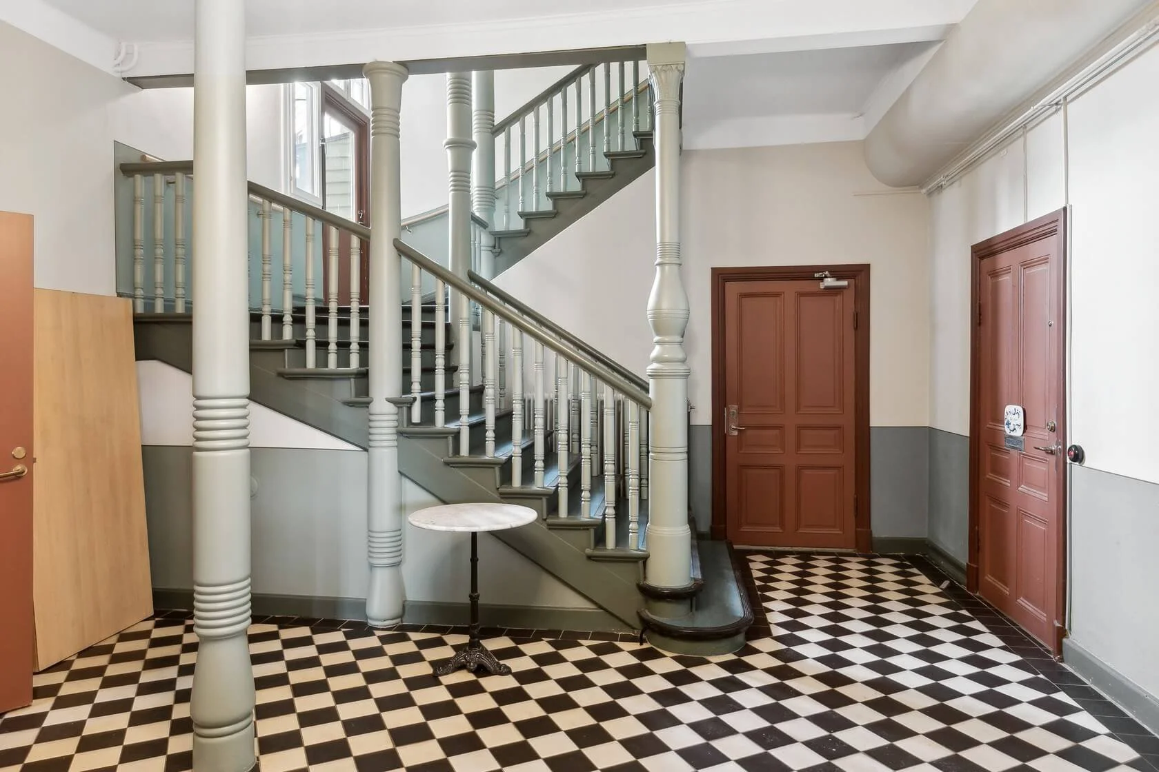 Interior of a building with a checkered black and white tile floor, a staircase with wooden railings, two brown doors, and a small round side table.