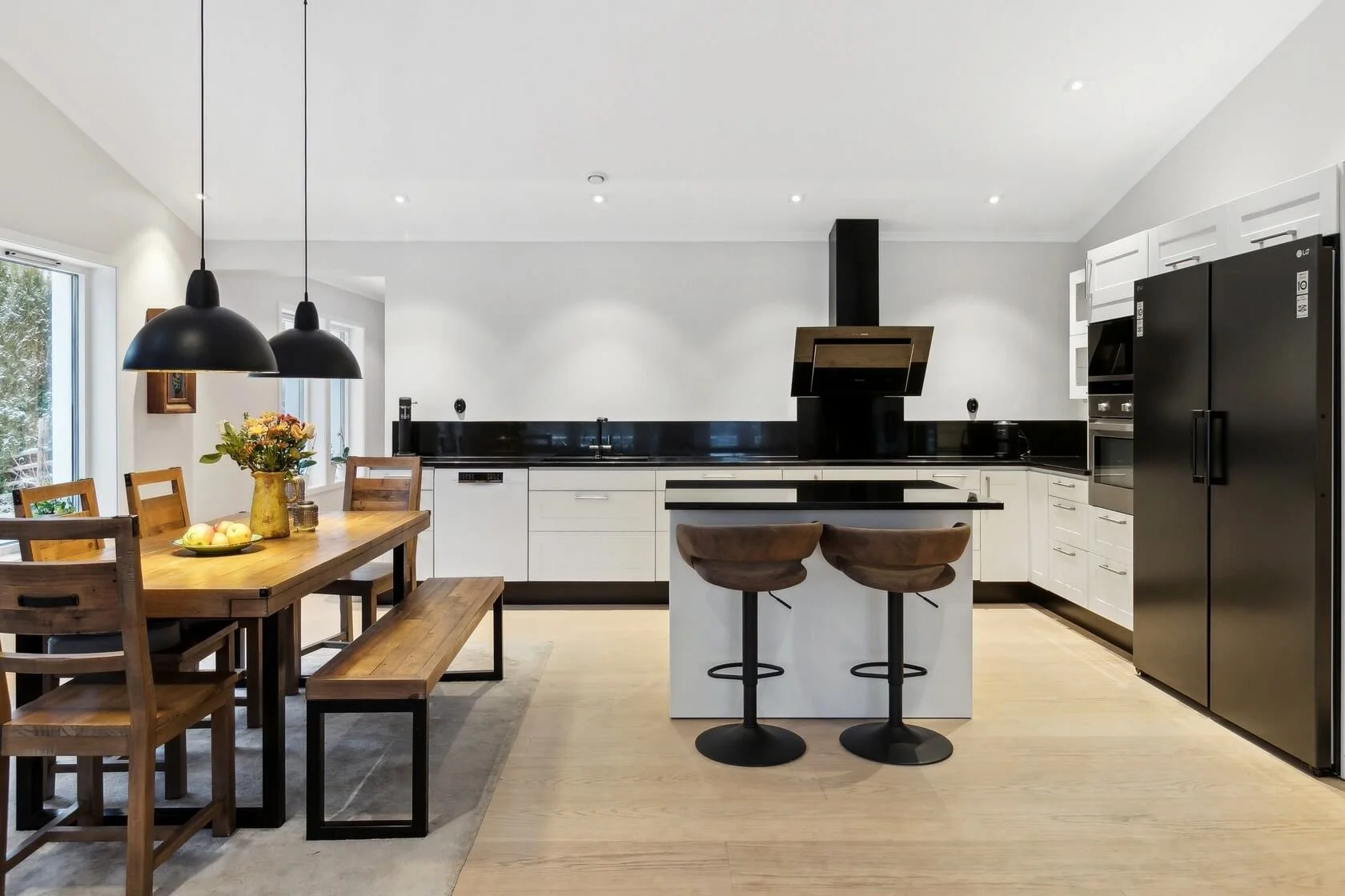 Modern kitchen with black and white cabinets, black refrigerator, black range hood, and a wooden dining table with chairs and a bench. There is a vase of flowers and a fruit bowl on the table, and two bar stools at the kitchen island.