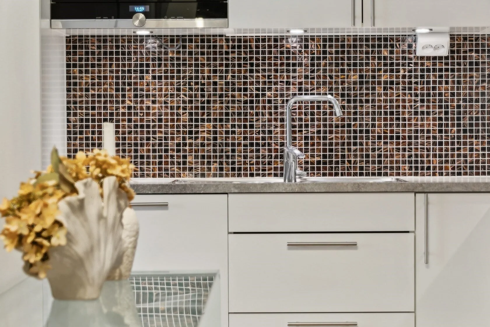 Modern kitchen with white cabinets, a stainless steel faucet, and a mosaic tile backsplash in shades of brown. There is a vase with dried flowers on the counter and a digital clock display on an oven above.