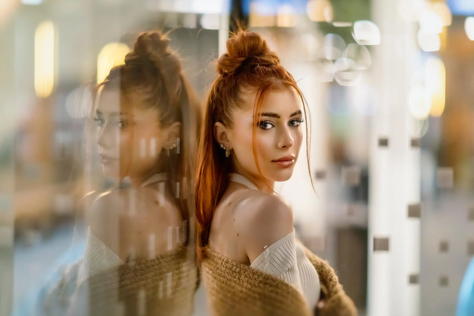 A young woman with red hair in a half-up style, wearing a white off-shoulder top and a brown cardigan, stands beside a reflective glass window, with city lights blurred in the background.