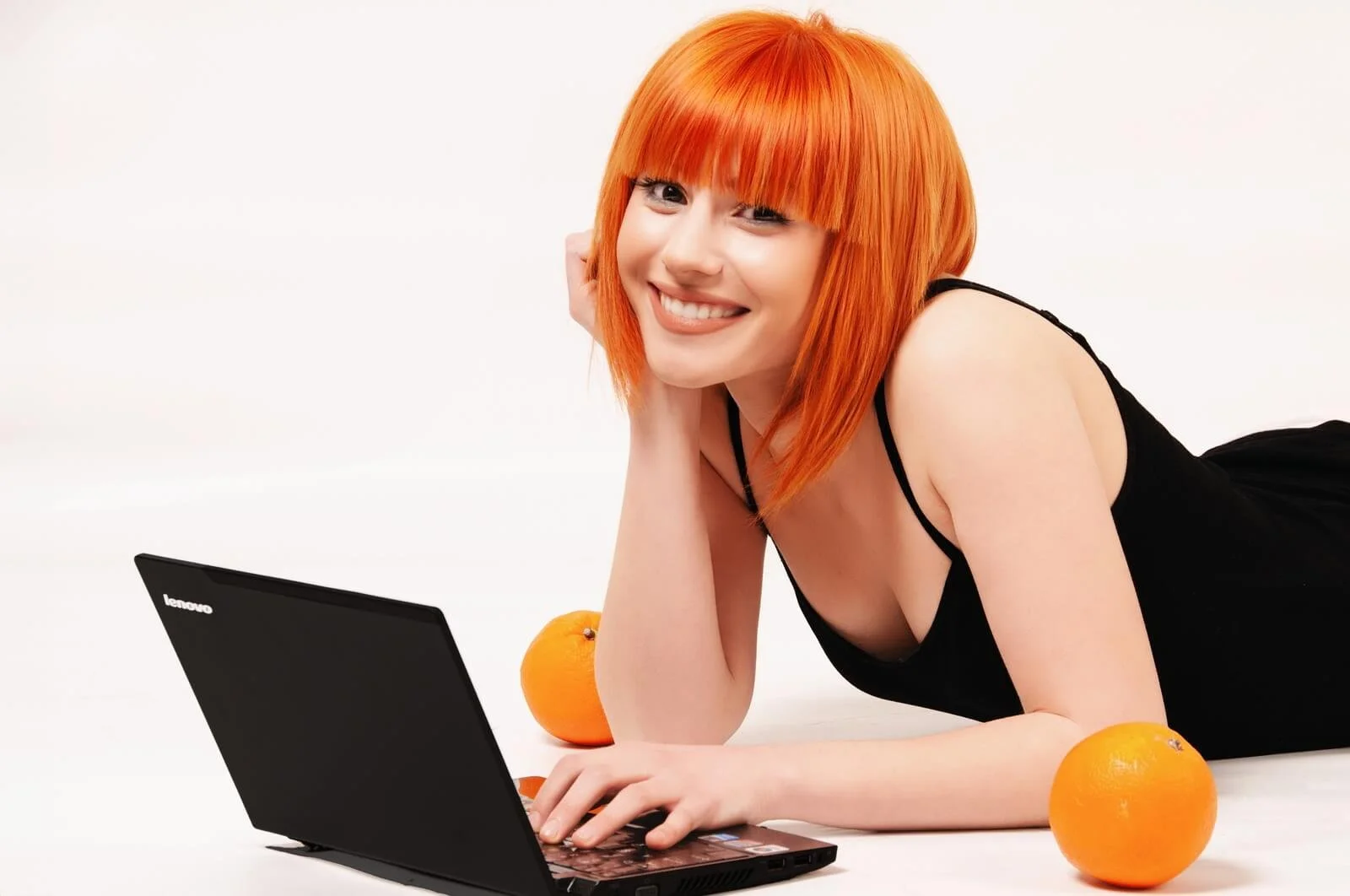 A smiling woman with bright orange hair lying on her stomach on a white surface, wearing a black dress, with a black laptop and oranges around her.