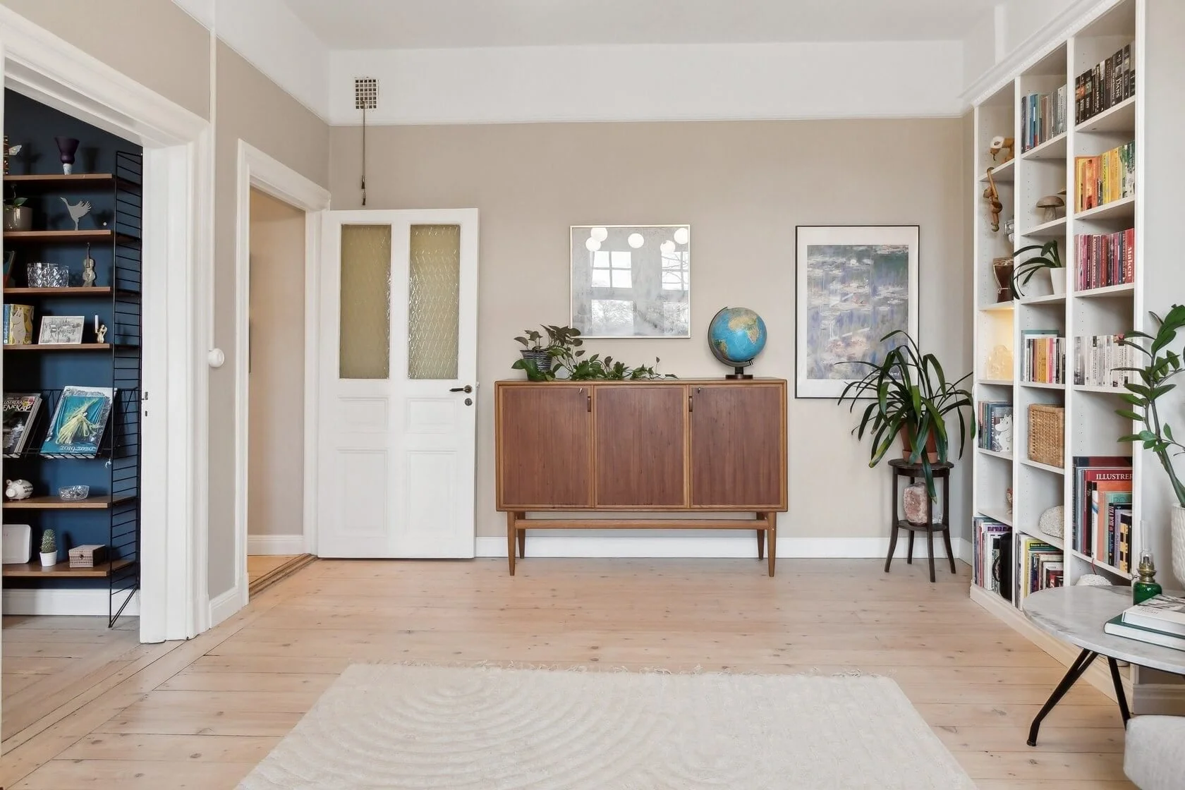 A living room with light hardwood flooring, a white bookshelf filled with books and décor on the right, a wooden sideboard with plants and a globe in the center of the back wall, and a small round table with books and a plant on the right.