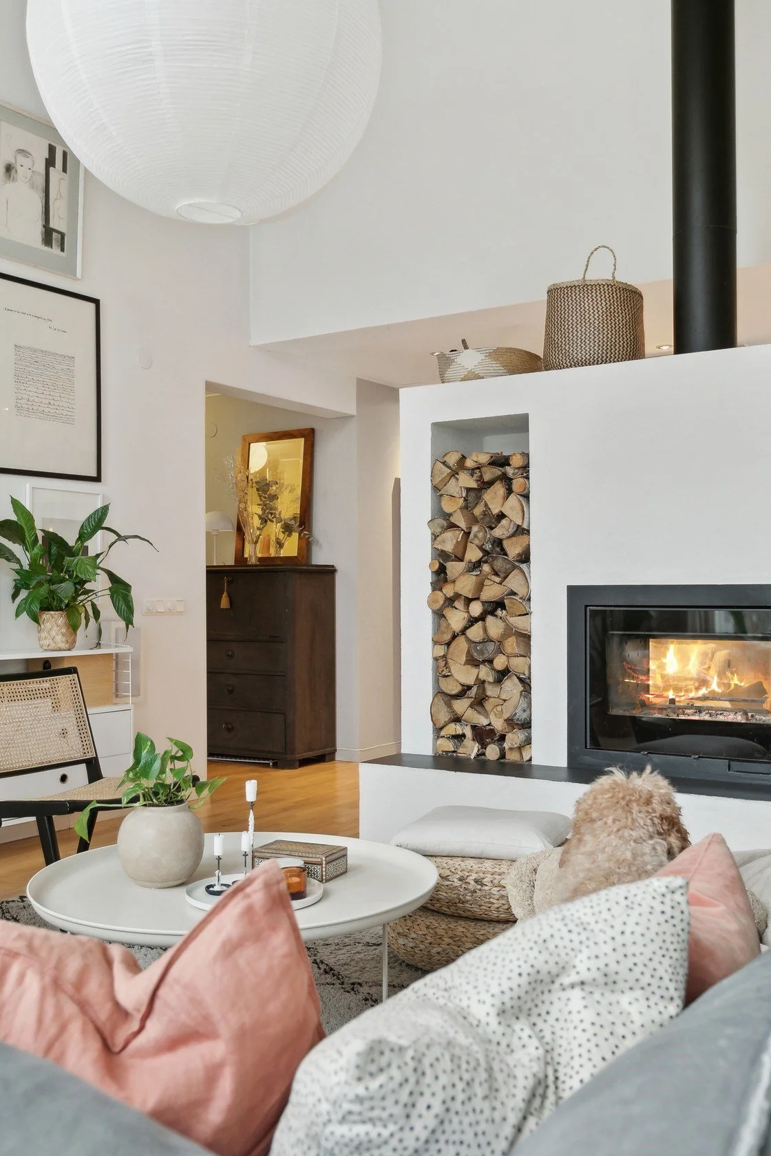 Living room with a fireplace, stacked firewood, a cozy dog on the sofa, and decorative pillows and plants.