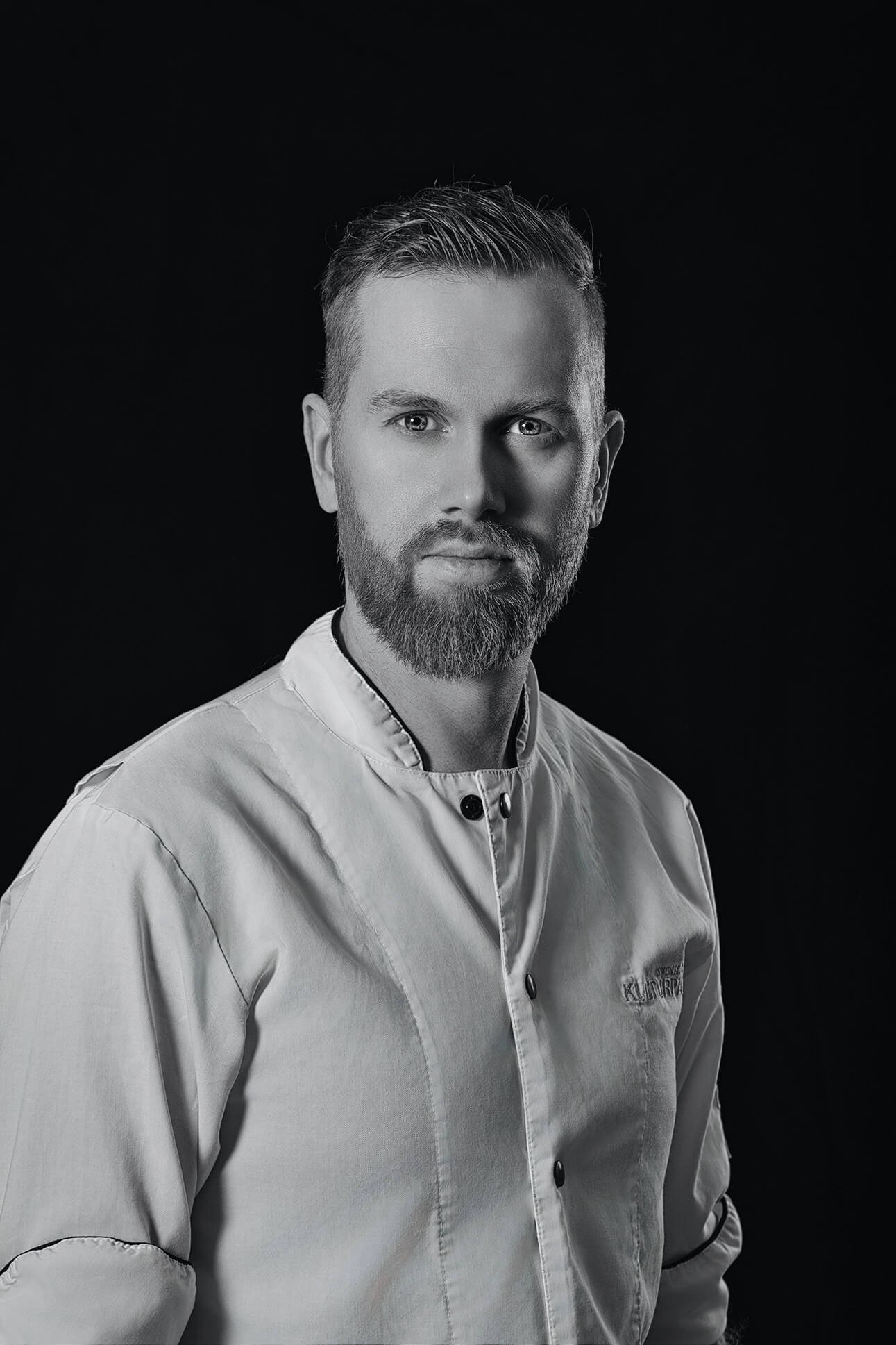 Black and white portrait of a man with a beard, wearing a chef's coat, against a solid dark background.