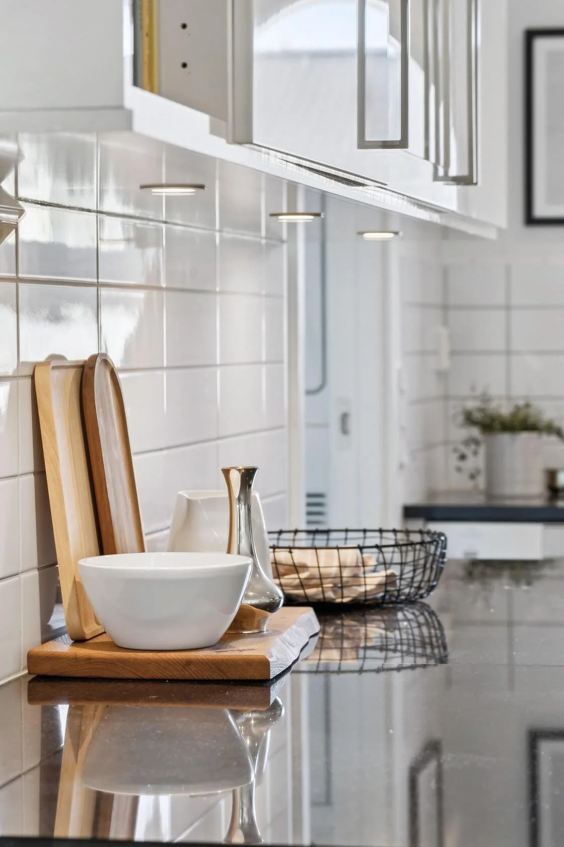 A modern kitchen countertop with white dishes, a wooden tray, and a wire basket, with white tiled walls and cabinets.