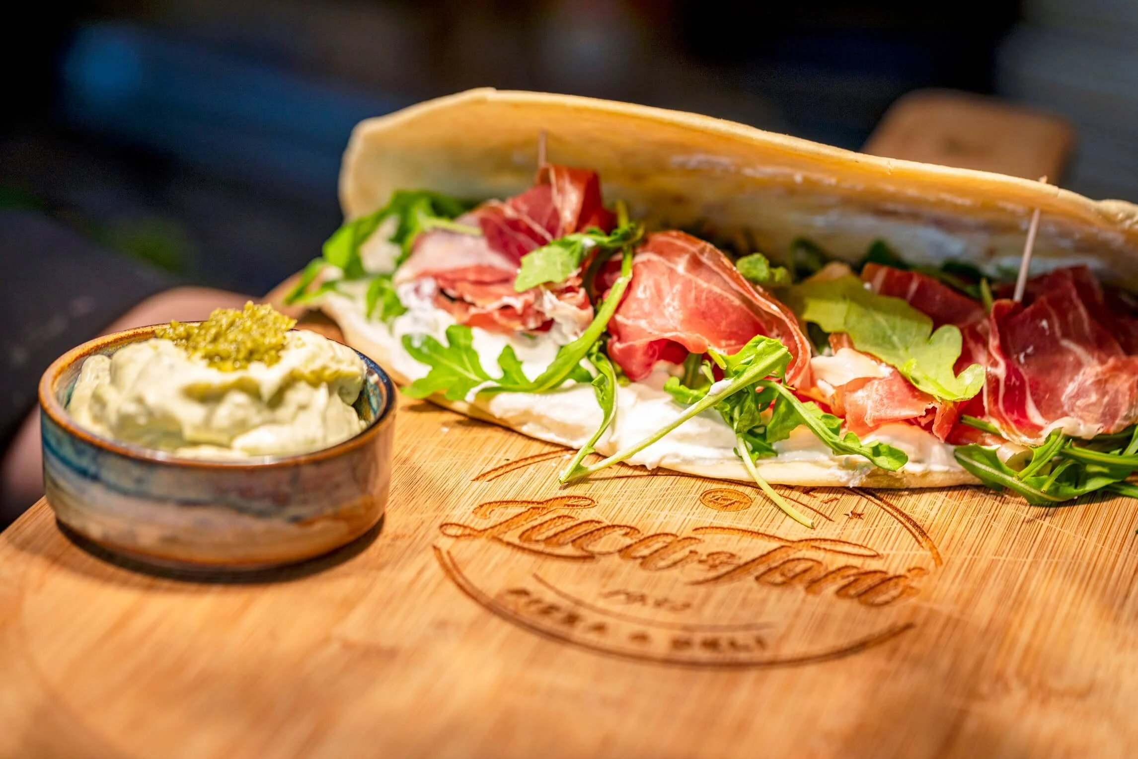 A close-up of a sandwich on a wooden board with green arugula, slices of prosciutto, and cheese inside a pita or flatbread, with a side of creamy dip garnished with herbs.