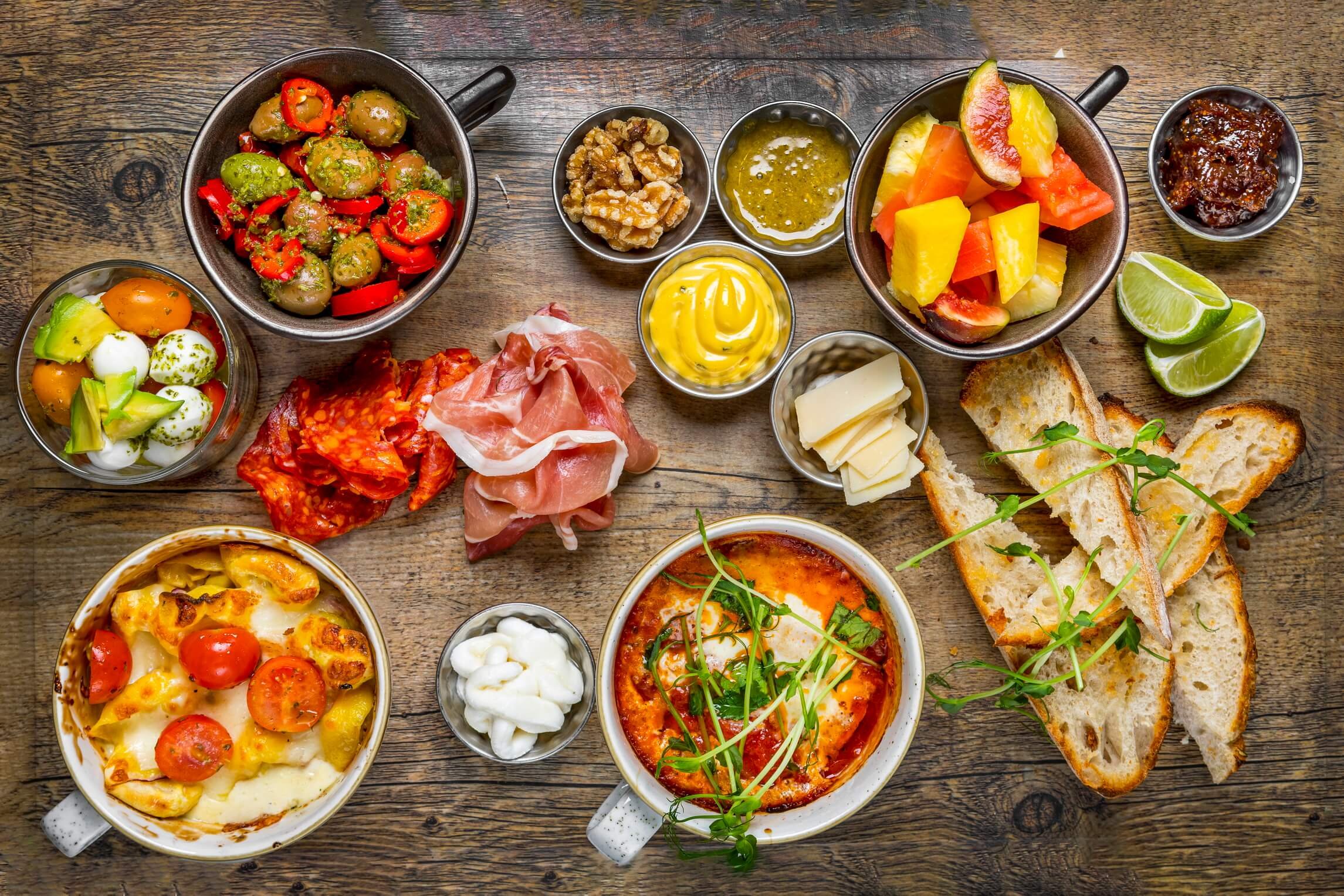 An assortment of tapas and appetizers on a wooden table, including marinated olives, sliced cured meats, cheese, bread, a baked dish with cherry tomatoes, and various bowls of dips, spreads, and fruits.