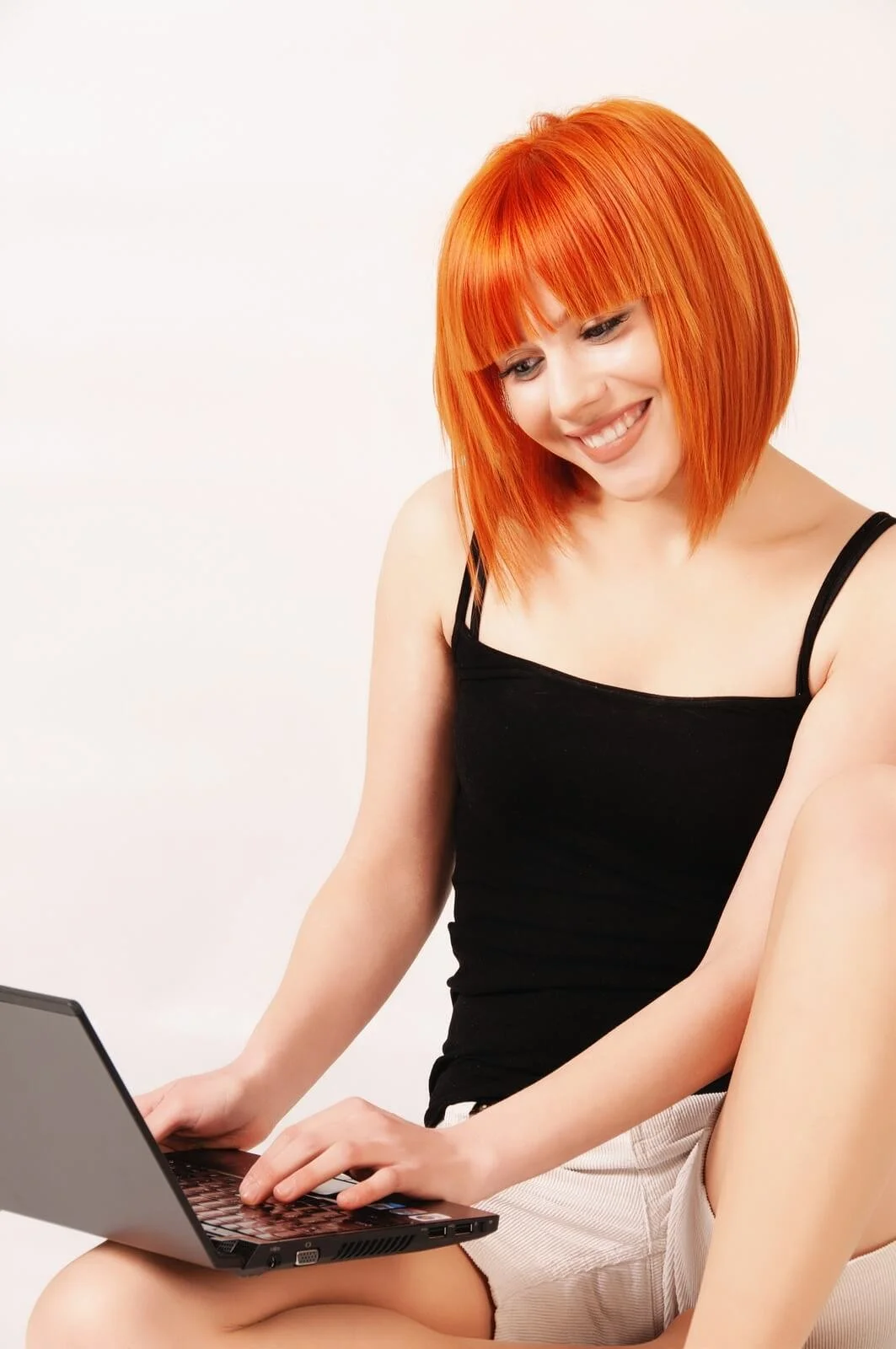 A young woman with bright orange hair, wearing a black sleeveless top and light-colored shorts, sitting cross-legged and using a laptop against a plain white background.