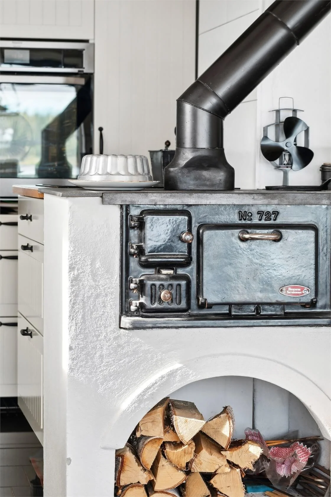 Close-up of a vintage black cast iron wood stove with a metal pipe vent, a metal door, and a small window, under a countertop with a dish covered by a metal lid, in a kitchen with white cabinetry and a window in the background. Wood logs are stored u
