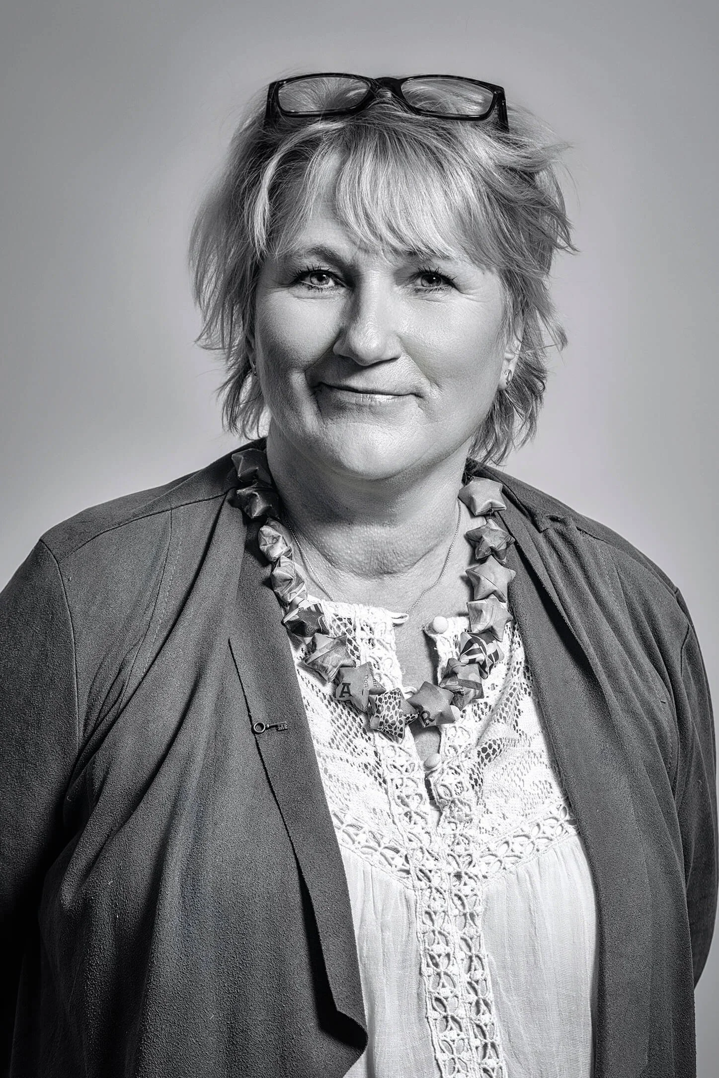 Black and white portrait of a middle-aged woman with short, wavy hair, wearing glasses on her head, a patterned blouse, a necklace, and a blazer.