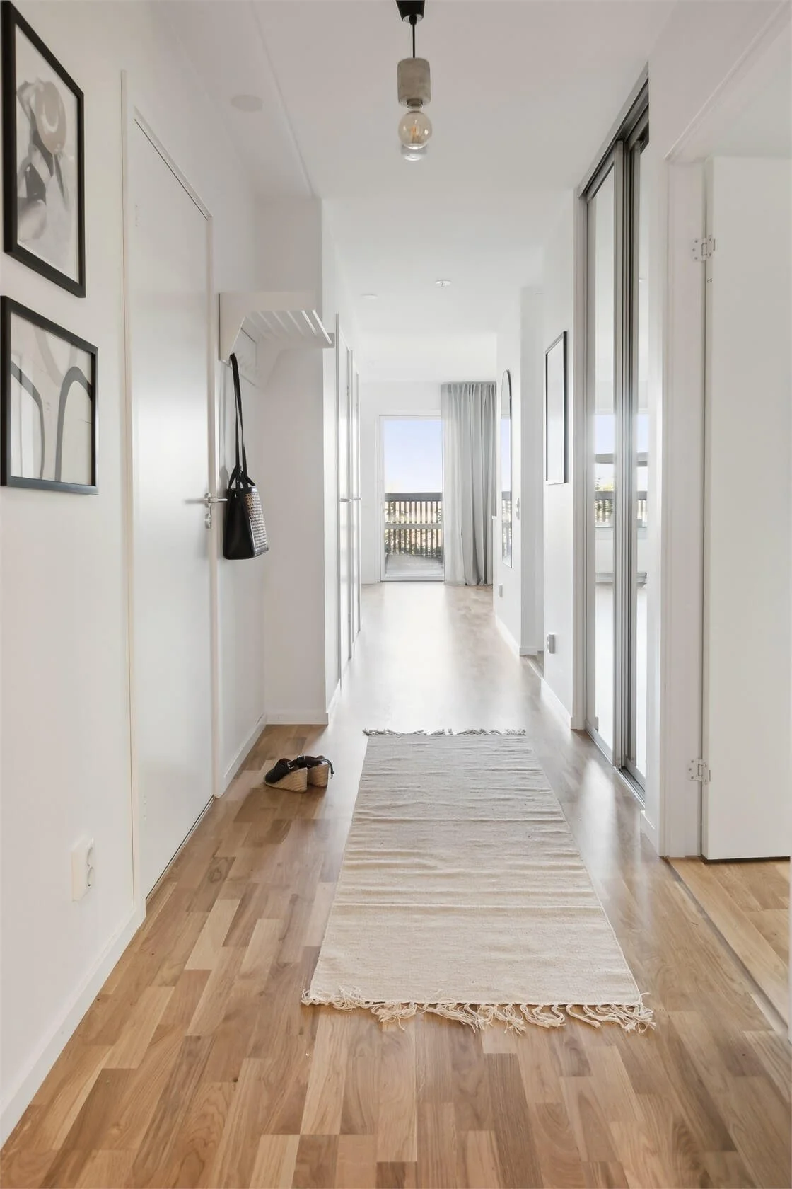 Bright hallway with light wood flooring, a beige runner rug, and a door on the left with framed artwork. Natural light from a balcony with sliding glass doors at the end, which are partially open with a curtain on one side. Shoes are on the floor nea