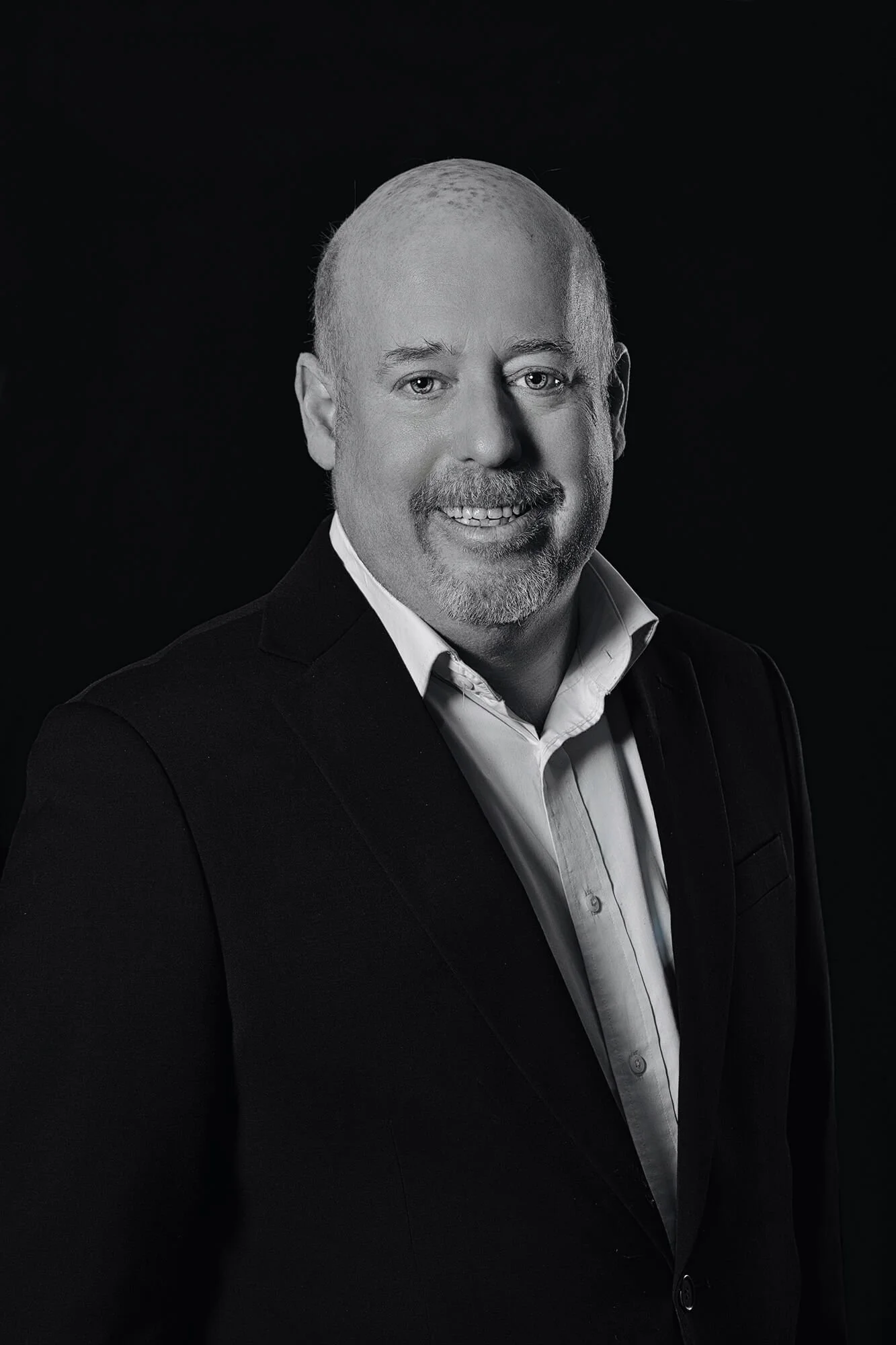 Black and white portrait of a smiling middle-aged man with a beard, wearing a dark blazer and a light-colored dress shirt, against a dark background.