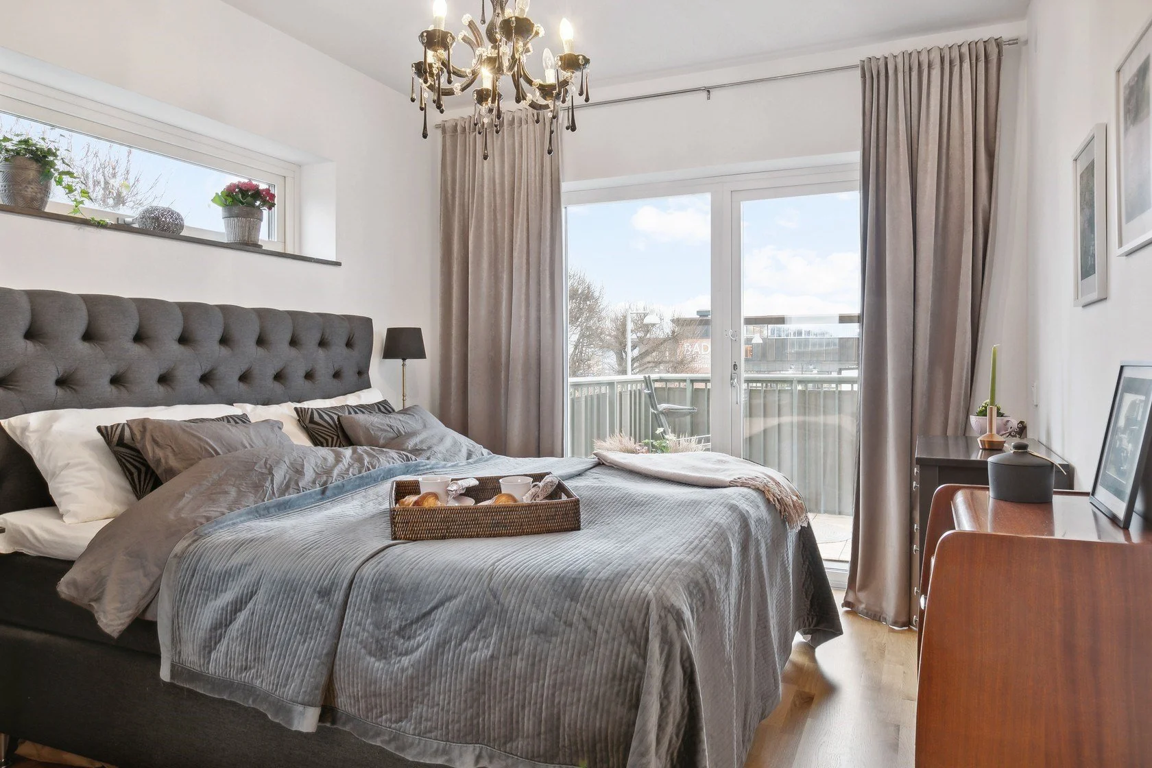 A bedroom with a large bed, gray bedding, and several pillows, near sliding glass doors leading to a balcony. The room features a black bedside lamp, a black chandelier, and a wooden dresser with a framed picture, a candle, and decorative items.