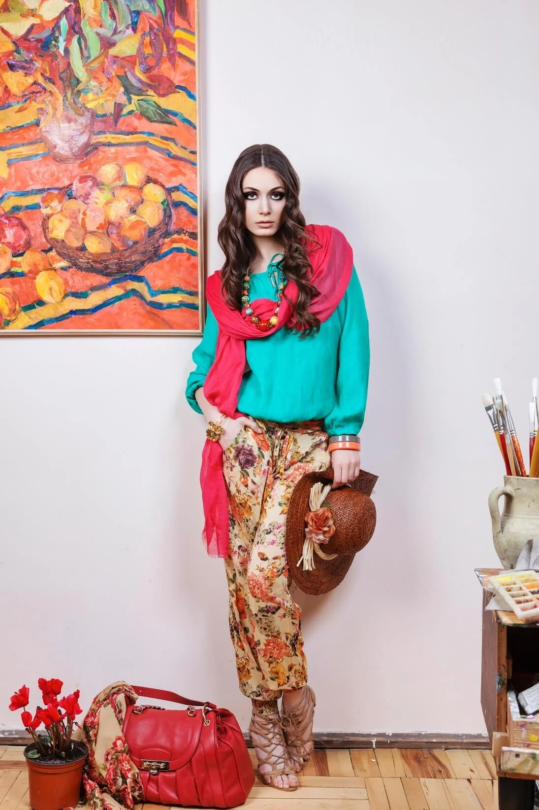 A woman standing indoors with an abstract floral painting behind her. She is wearing a teal top, floral pants, a red scarf, and colorful accessories. She holds a straw hat and is surrounded by a red bag, a small potted plant with red flowers, and art