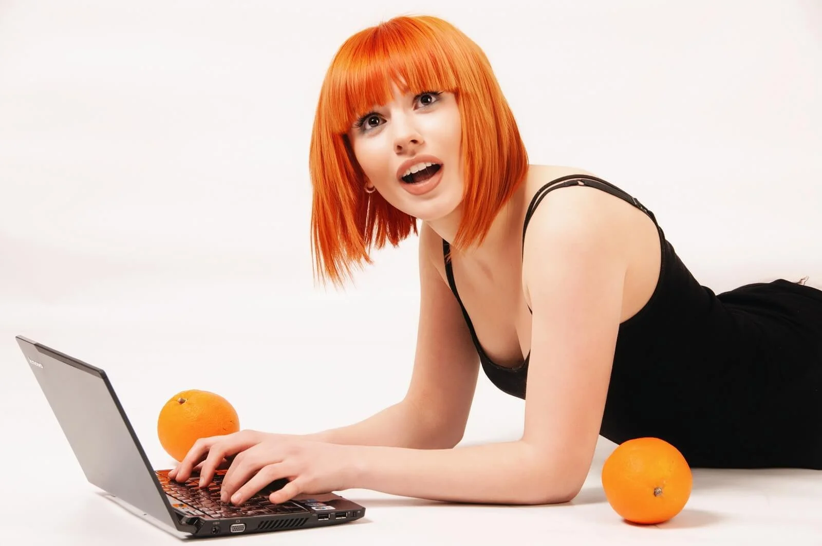 A woman with vibrant red hair, wearing a black tank top, lying on her stomach on a white floor, using a laptop with two oranges beside her. She has an expression of surprise or curiosity.
