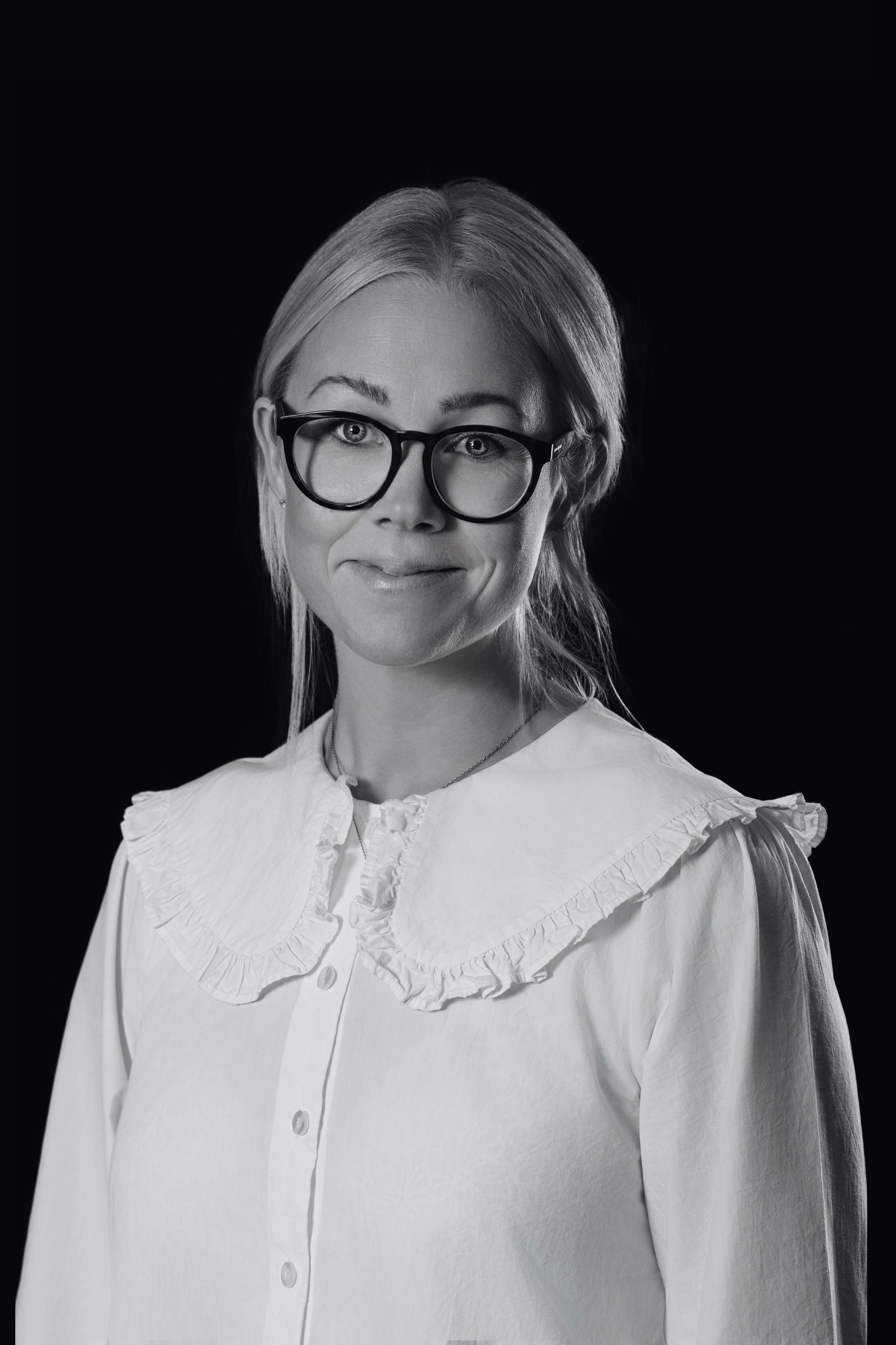 Black and white portrait of a woman with glasses, wearing a white blouse with a ruffled collar, smiling softly against a dark background.