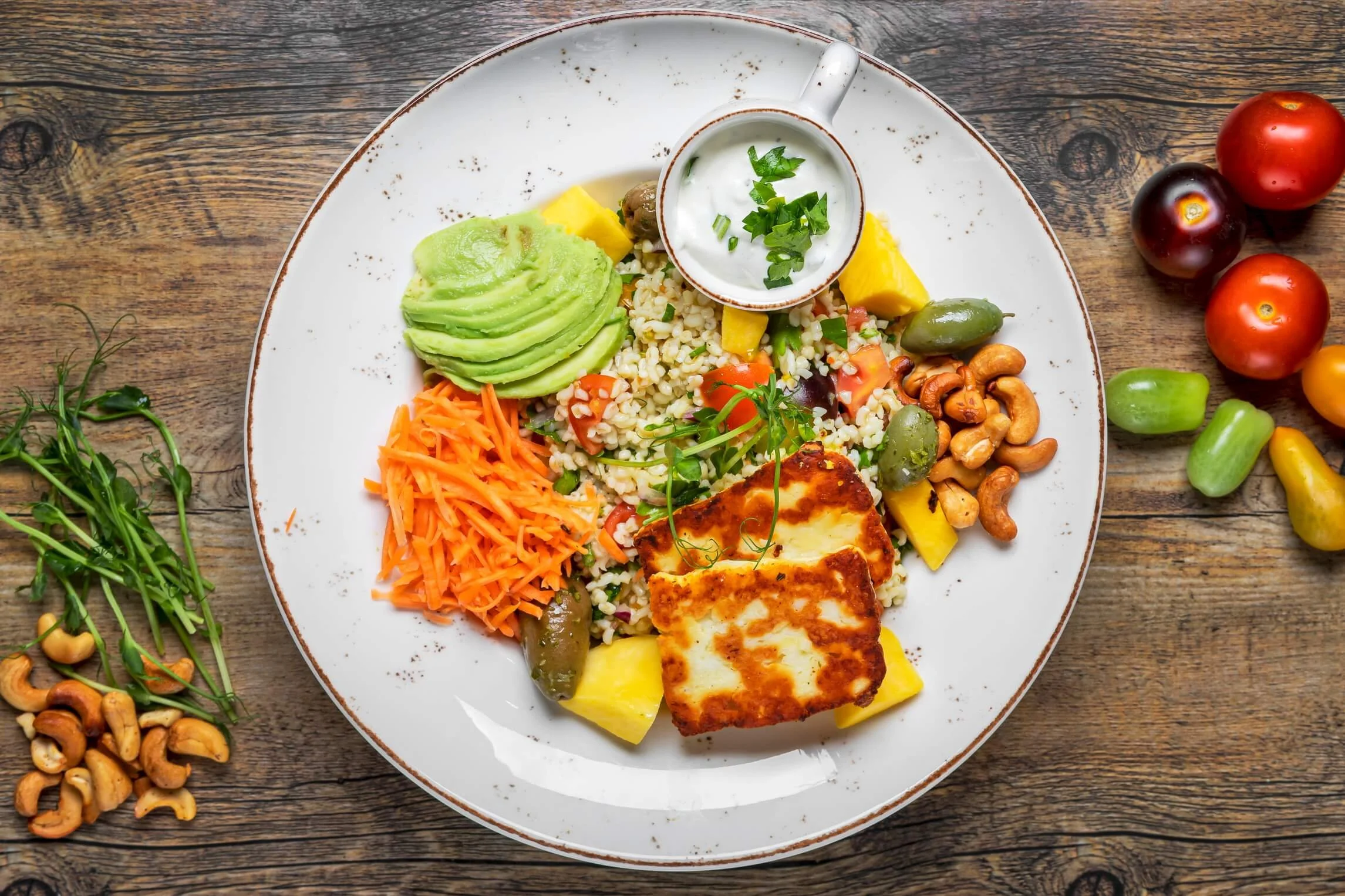 Plate of grilled tofu with yellow mango slices, sliced avocado, shredded carrots, couscous with vegetables, green olives, cashews, and a small bowl of yogurt sauce garnished with chopped herbs on a wooden table with tomatoes and green grapes nearby.
