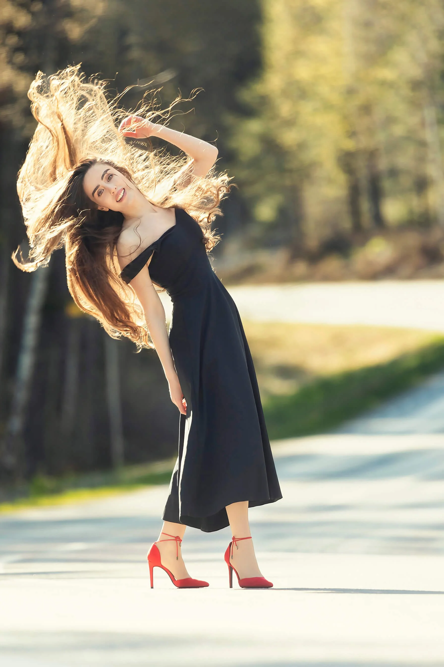 A woman with long, wavy brown hair, wearing a black dress and red high heels, is striking a playful pose outdoors during daytime, with trees and sunlight in the background.