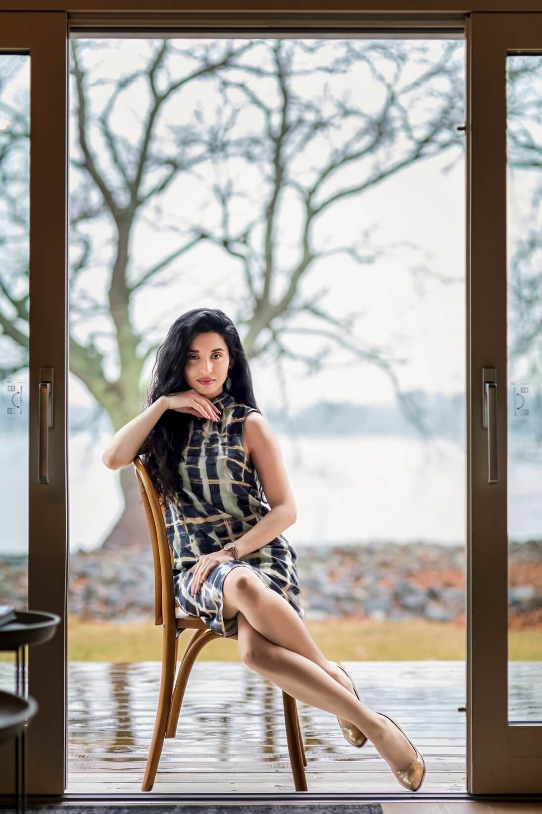 A woman sitting on a wooden chair in front of a large glass door with a view of trees and a lake.