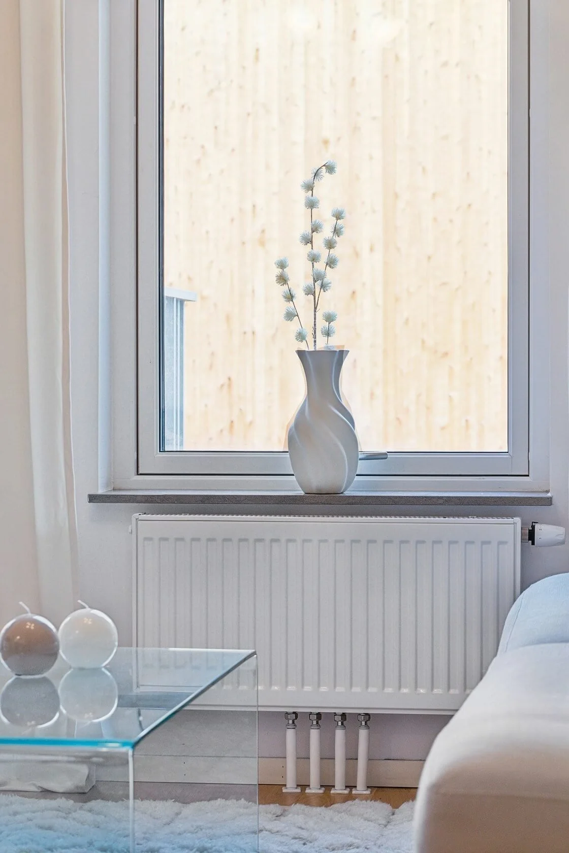A white vase with a curvy shape and vertical stripes holding tall, fluffy, light blue flowers sits on a windowsill in a bright, modern living room. The window shows a wooden fence outside. In front of the window, there's a white radiator and a glass 