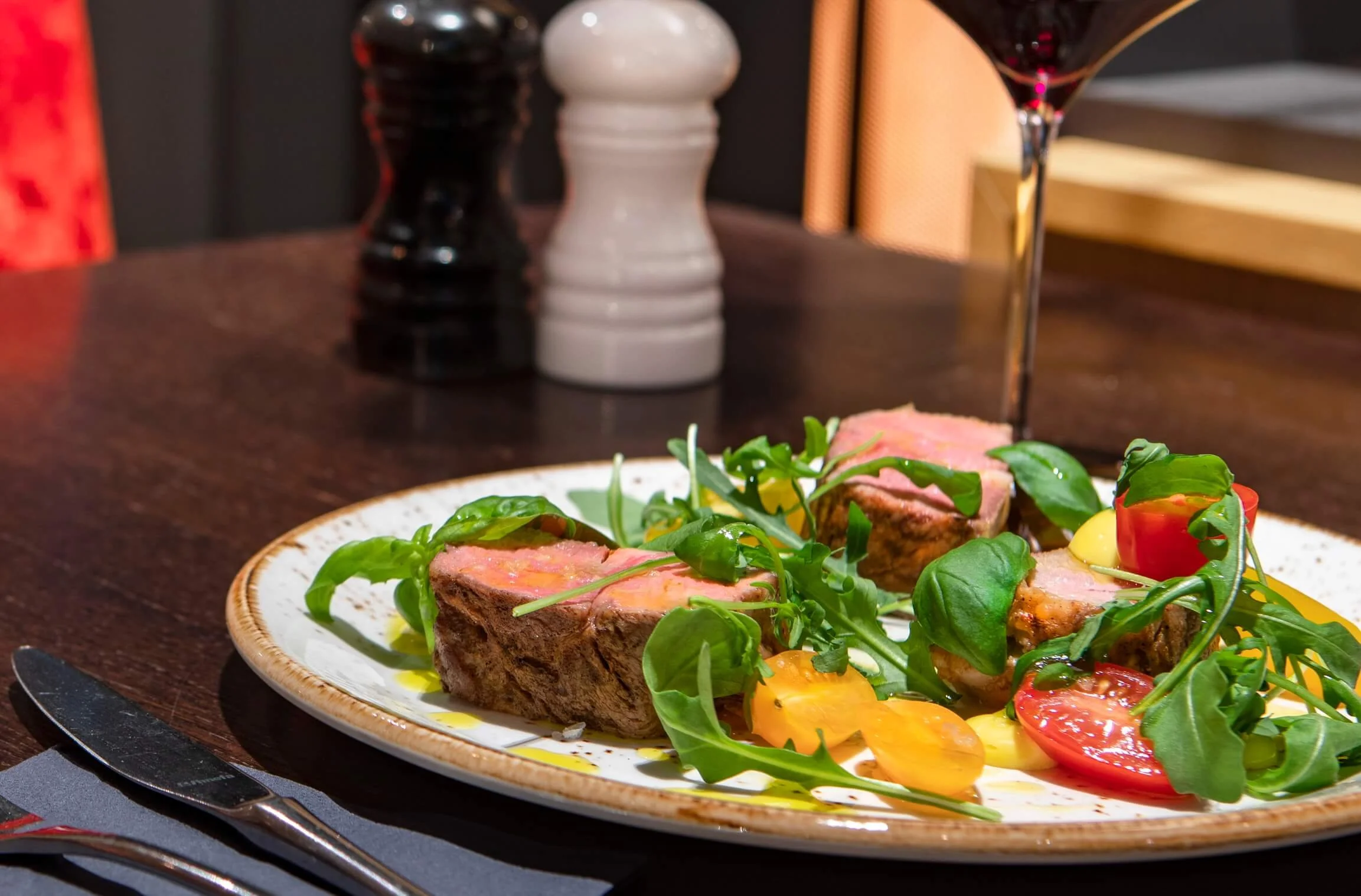 A plate with seared tuna topped with slices of pink tuna, surrounded by fresh green salad leaves, cherry tomato halves, and a yellow sauce. In the background, black and white salt and pepper shakers and a glass of red wine are visible on a dark woode