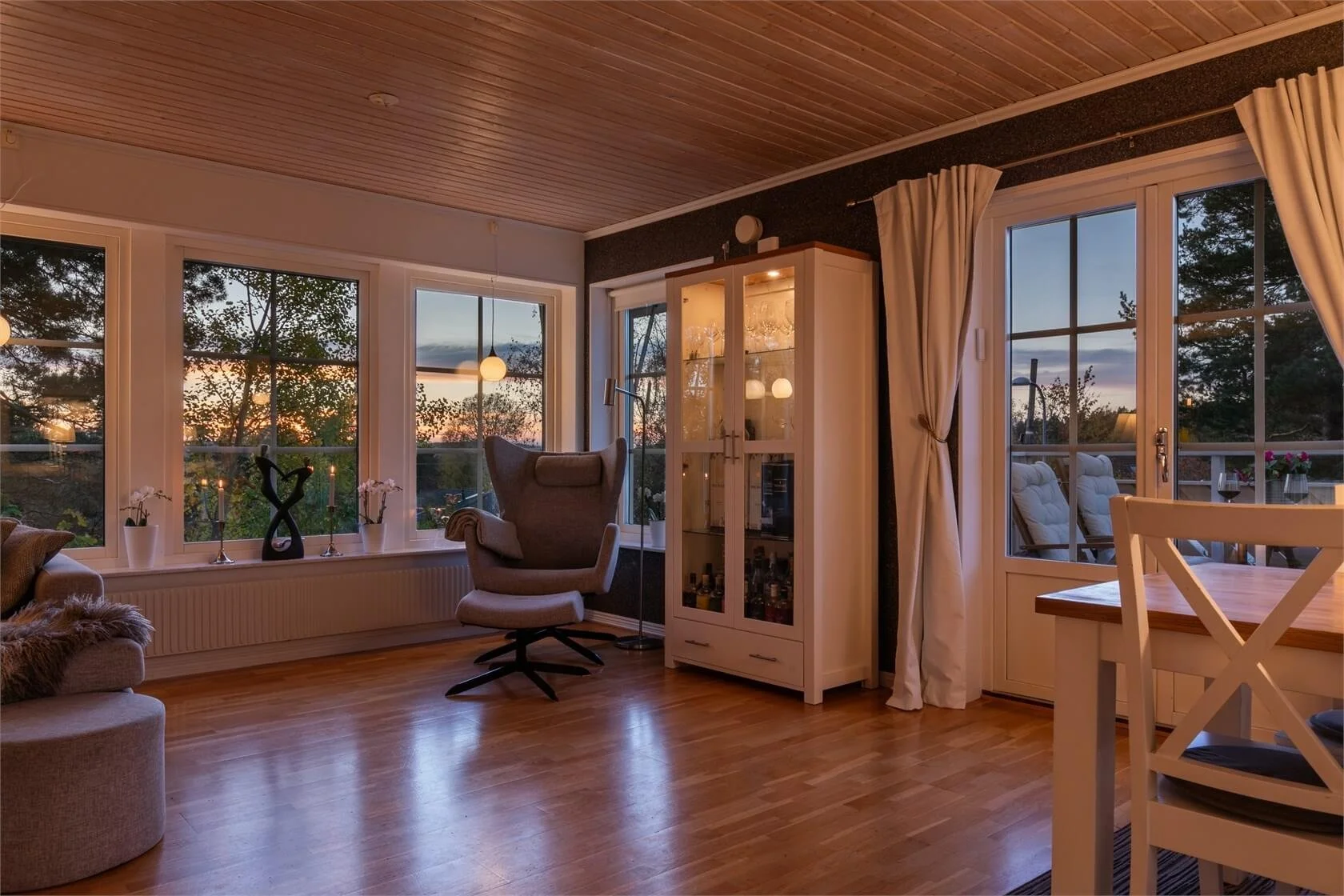Living room with large windows showing sunset, wooden ceiling, hardwood floor, white cabinet, gray armchair, curtains, and outdoor seating