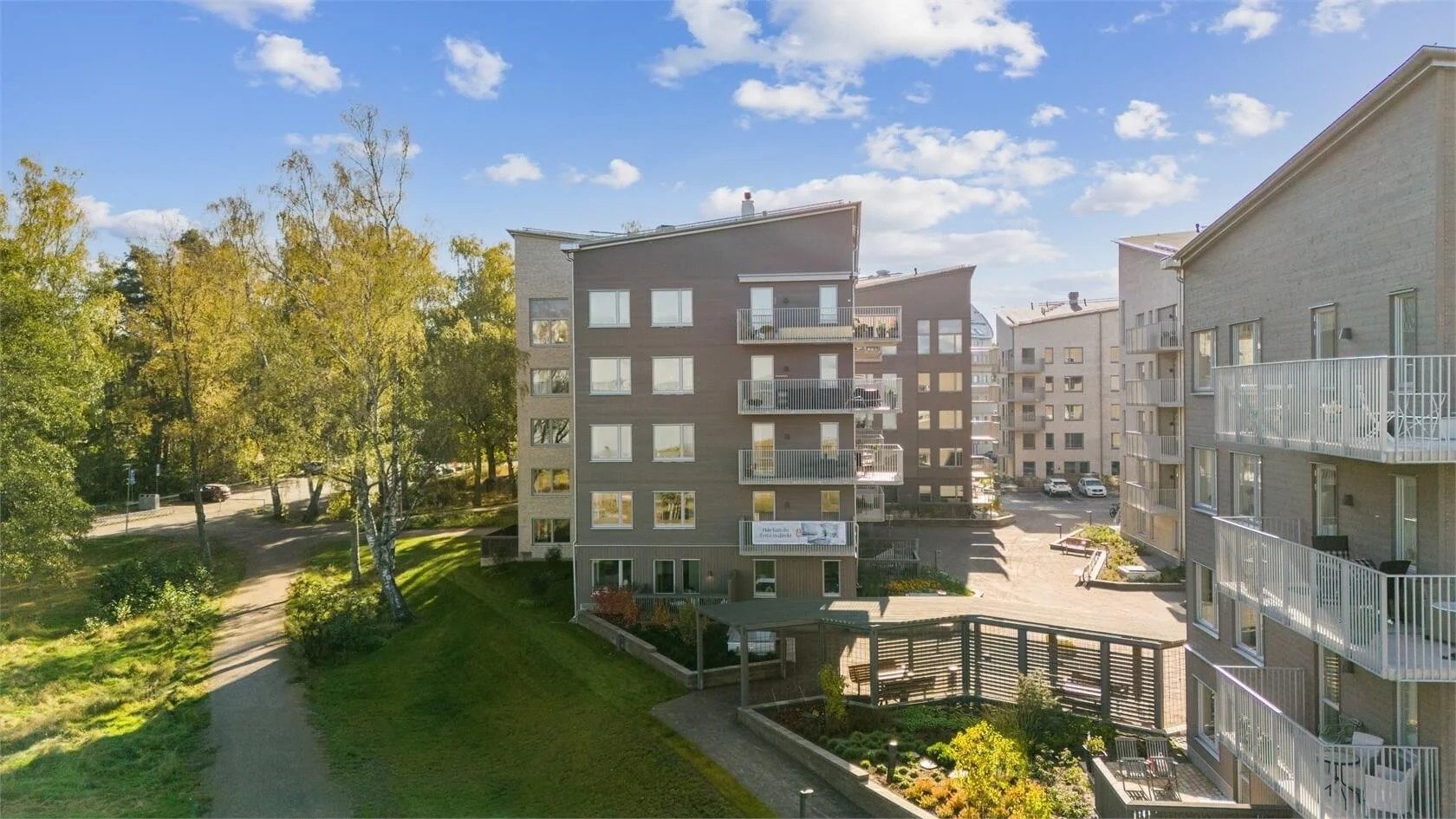 View of modern apartment buildings with balconies, surrounded by trees and greenery, under a partly cloudy sky.