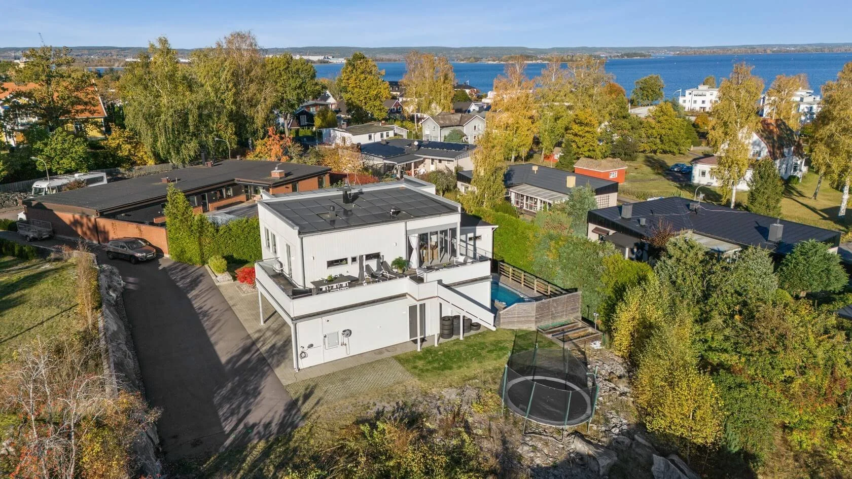 Aerial view of a modern white house with multiple outdoor patios, surrounded by trees with fall foliage, neighboring houses, a driveway with parked cars, a small swimming pool, and a trampoline near a rocky slope near a large body of water.