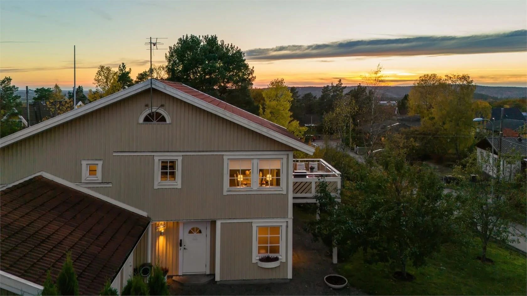 A house with beige siding, white trim, and a red-tiled roof, illuminated from inside at sunset, overlooking a neighborhood with trees and distant hills.