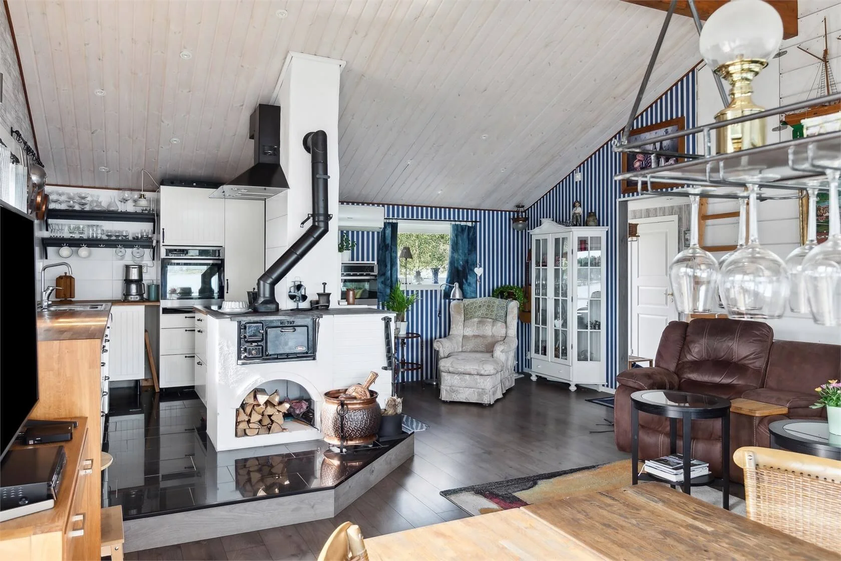 Living room with wood-paneled ceiling, striped blue wallpaper, standing armchair, brown sofa, glass cabinet, small tables, and wine glasses hanging from a rack.