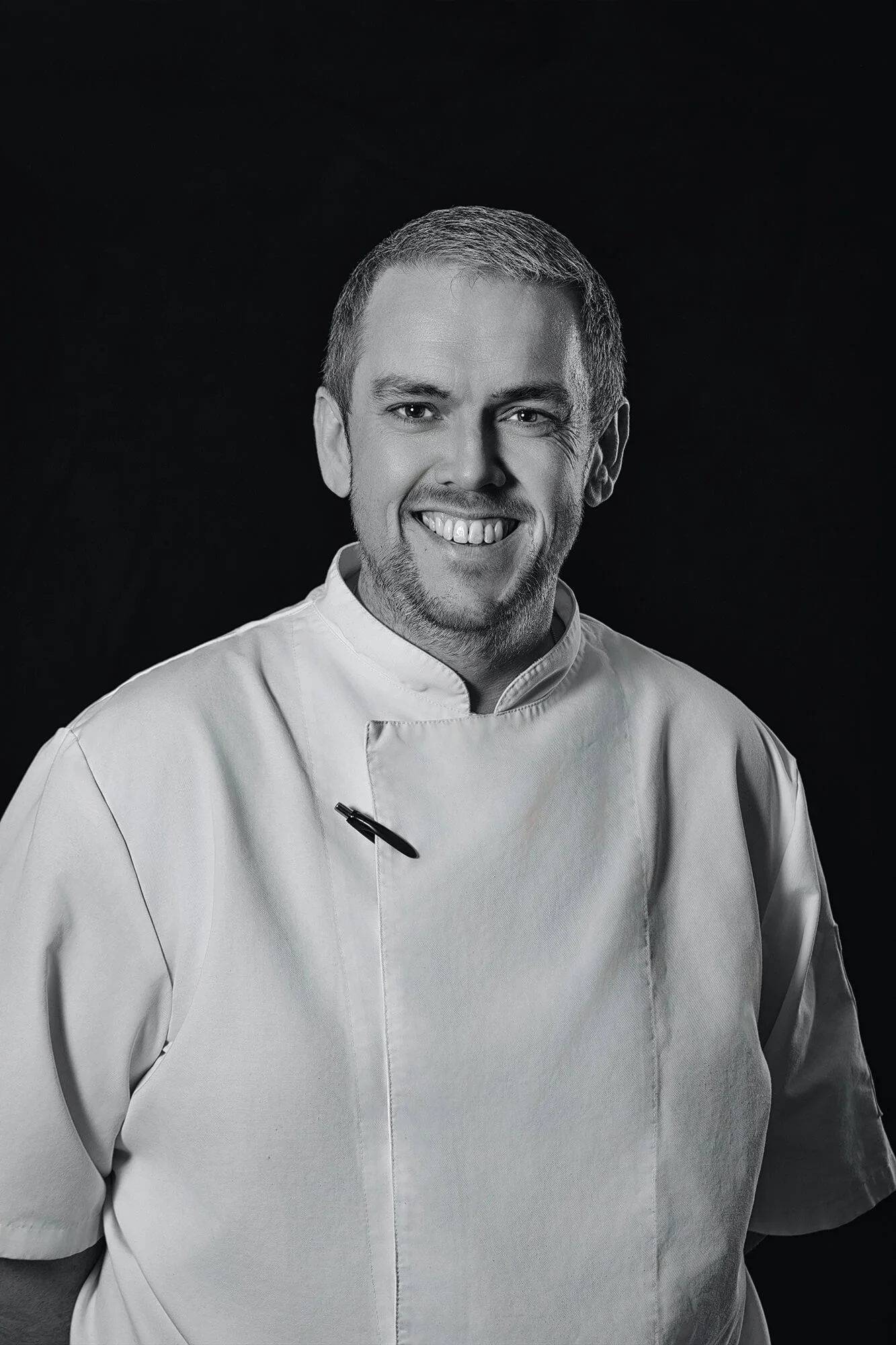 Black and white photo of a male chef in a white chef's coat smiling at the camera, dark background.