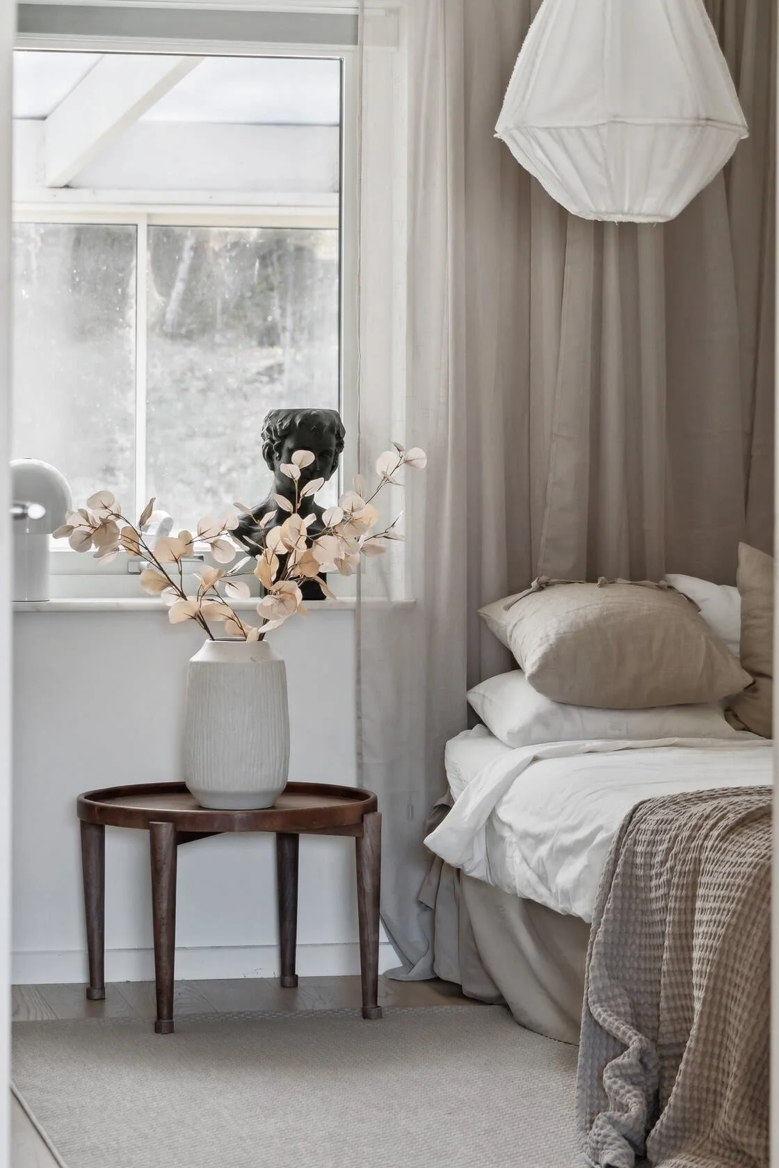 A bedroom corner with a bed, pillows, a wooden side table, a vase with flowers, a decorative bust, and curtains near a window.