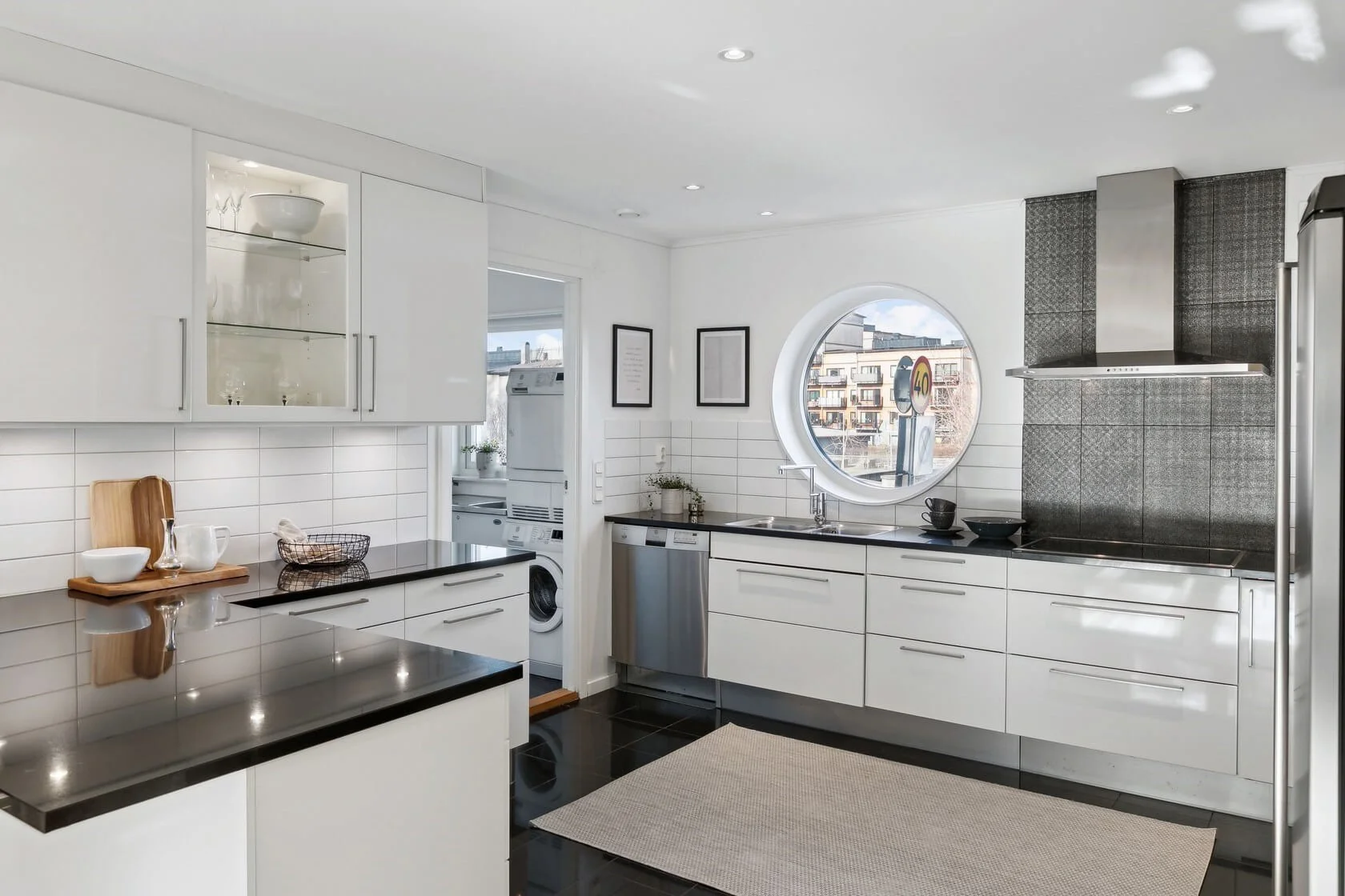 Modern kitchen with white cabinets, black countertops, a round window, and a view of neighboring buildings.