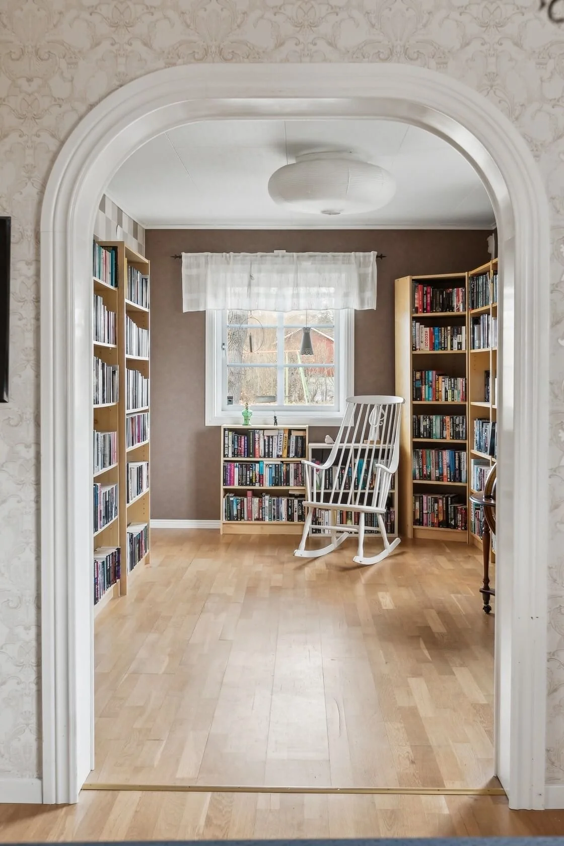 A cozy room with wooden bookshelves filled with books, a white rocking chair, a window with white curtains, a small shelf filled with books, and a light fixture on the ceiling.
